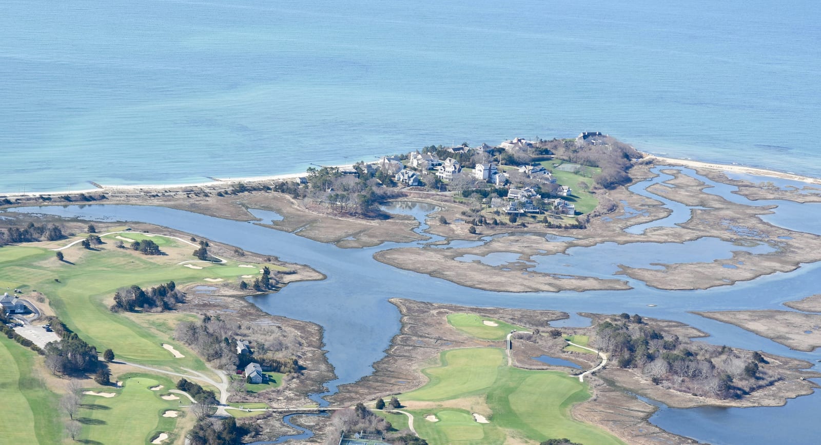 Aerial view of a coastal peninsula with marsh channels, a golf course, and homes along the shoreline.