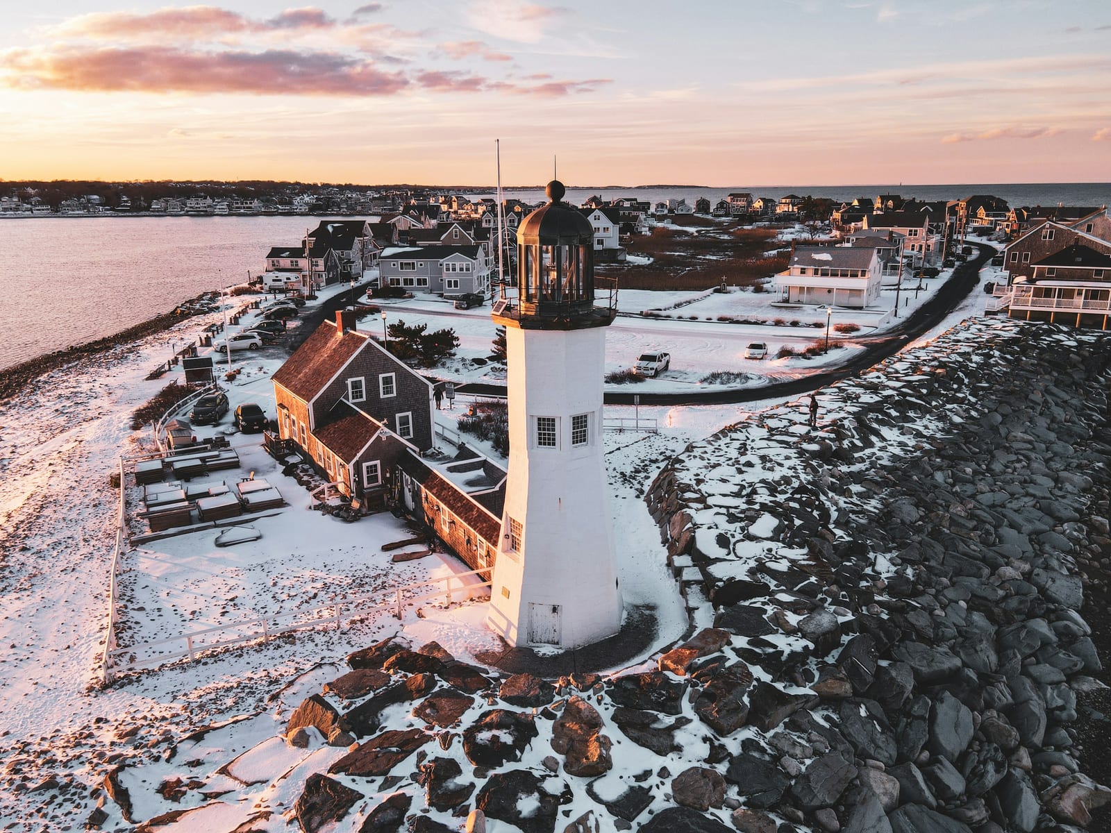 An aerial view of a snow-covered coastal town with a lighthouse lit by warm sunset light, waves lapping at a rocky point.
