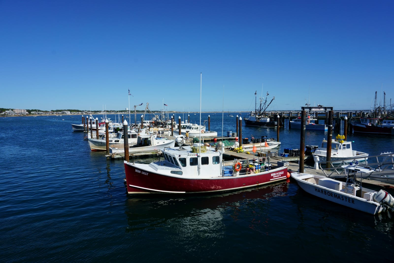 Red and white fishing boats tied up at a Cape Cod harbor pier under a clear blue sky.