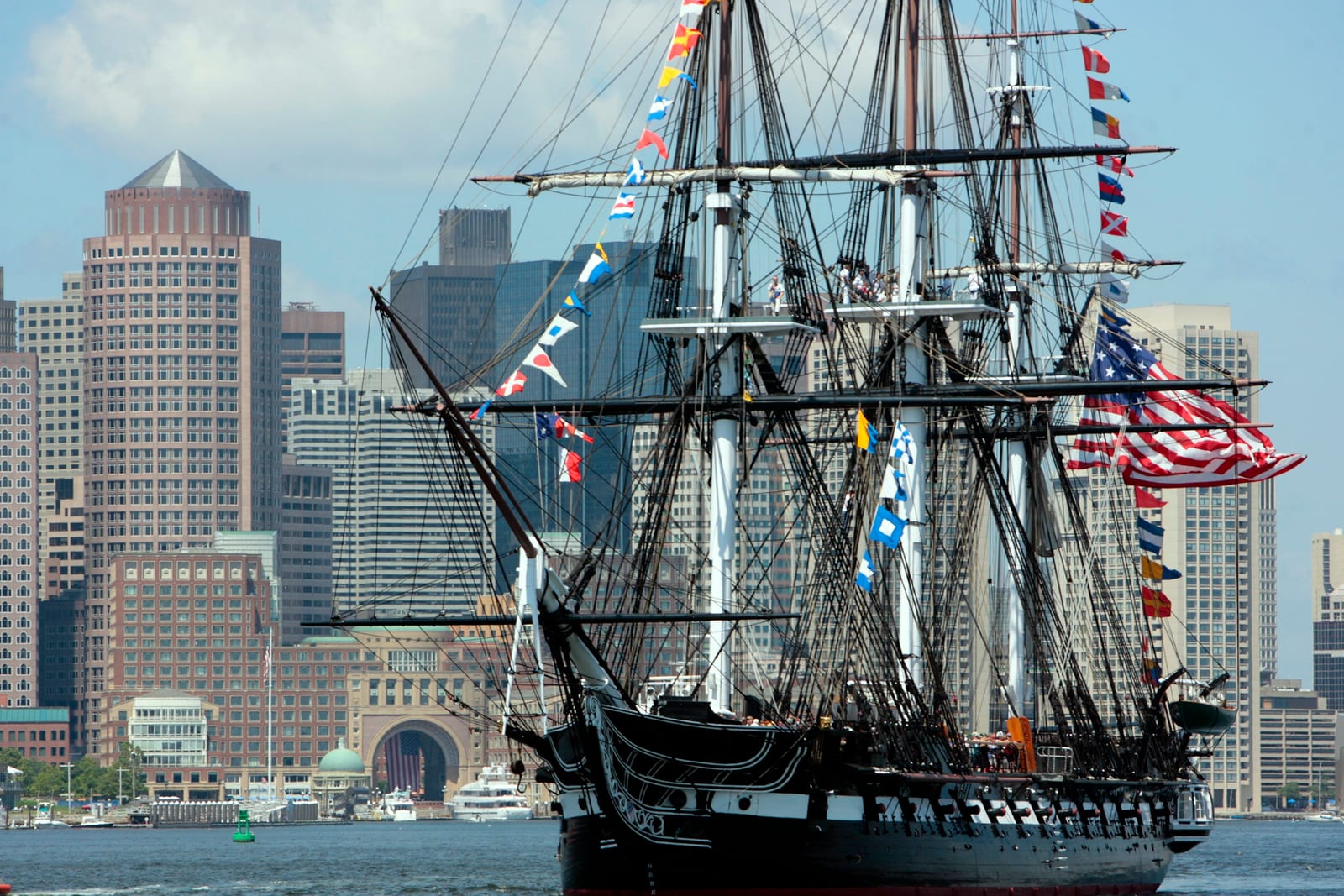 The USS Constitution, a three-masted wooden warship, under way on the water in Boston Harbor with the downtown skyline behind it