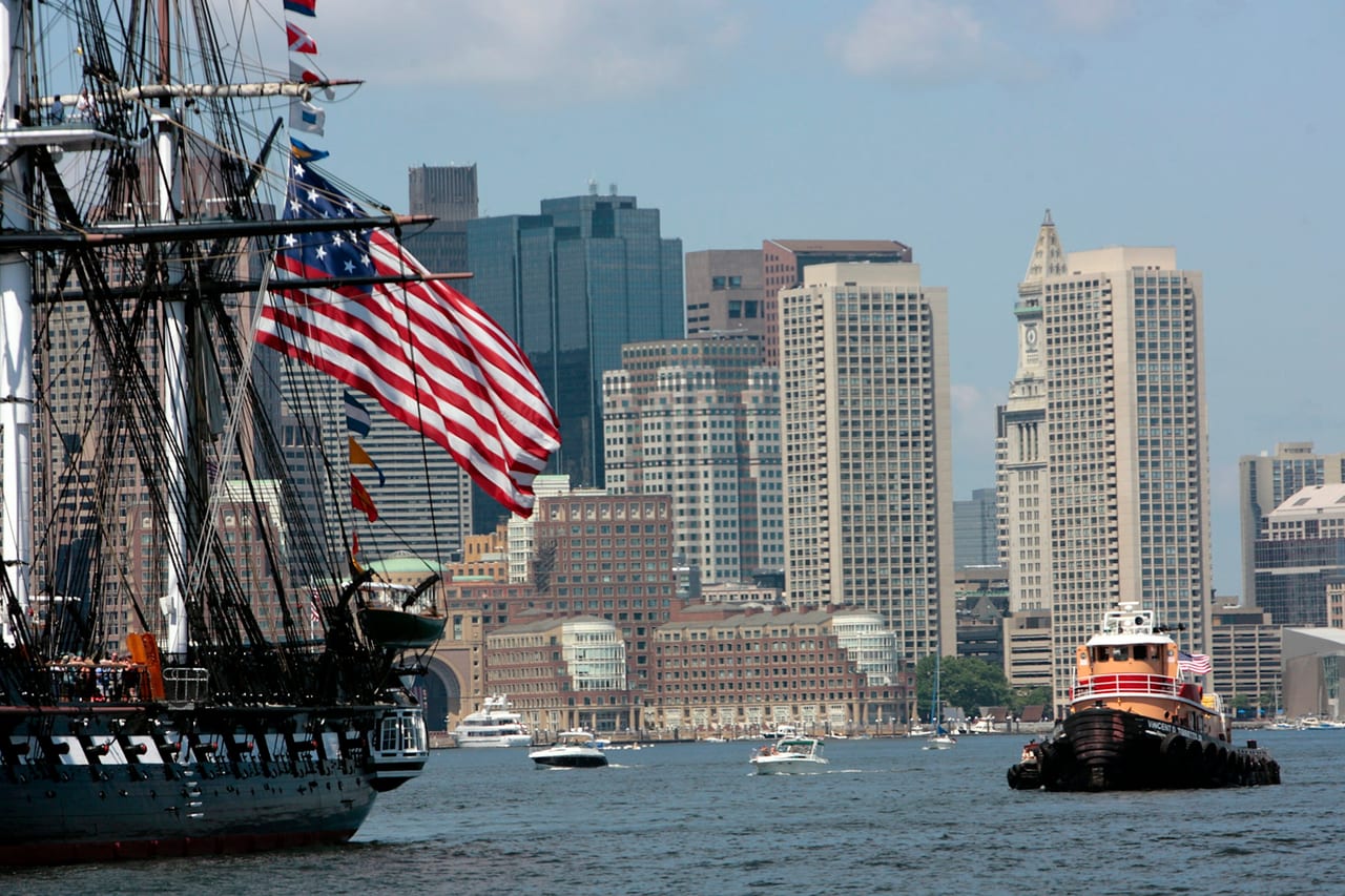 The USS Constitution sits at its berth in the Charlestown Navy Yard, its dark hull and tall masts reflected in the water.