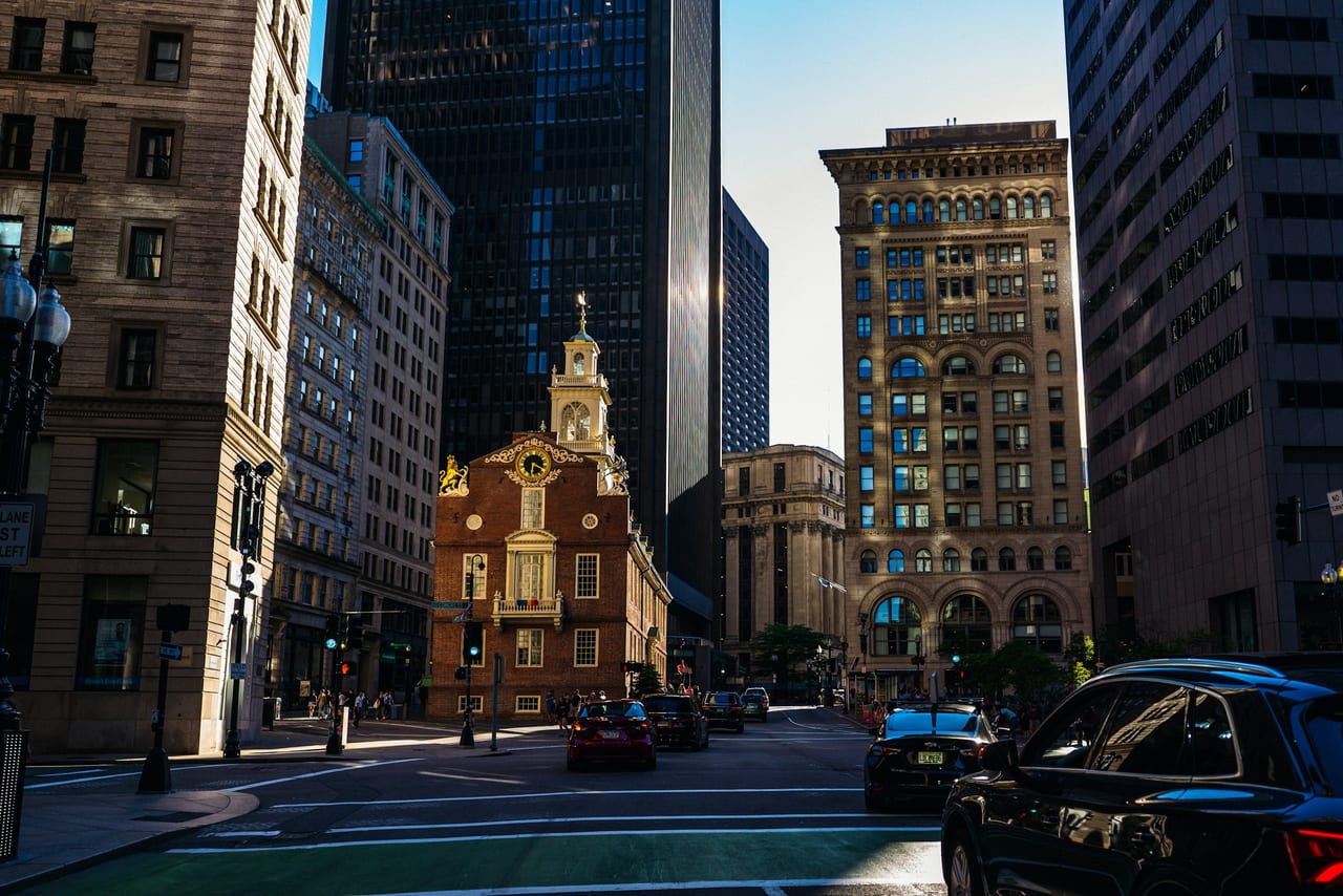 The red-brick Freedom Trail line crosses a downtown Boston sidewalk, with the Old State House in the distance.
