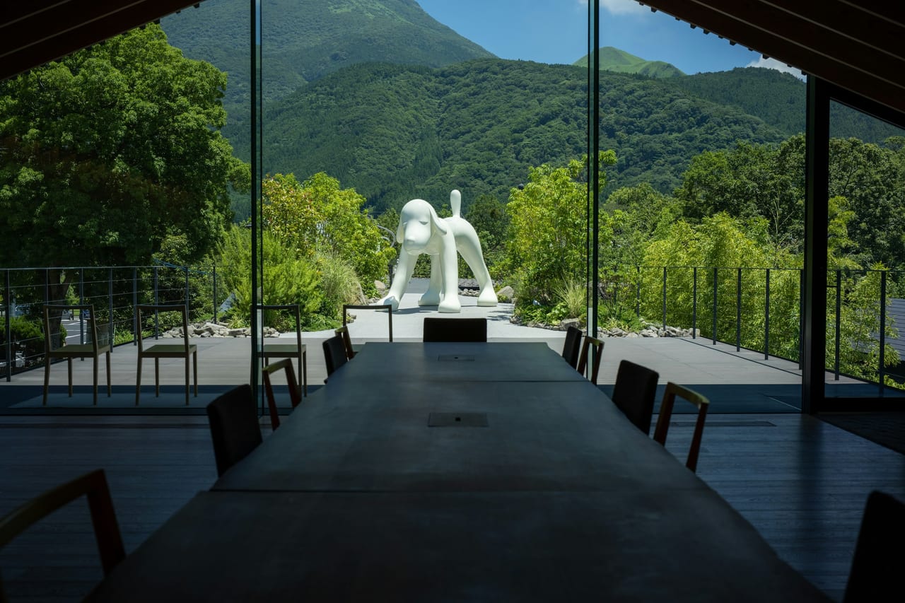 Bronze figures stand on a sunlit lawn before the museum's stained-glass Symphonic Sculpture tower, forested mountains behind.