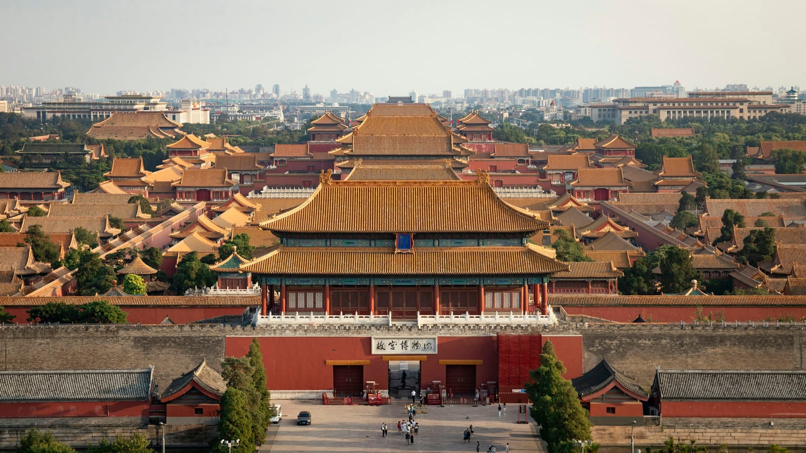 A bright elevated view over the Forbidden City palace complex, with red halls and golden roofs leading toward the city skyline.