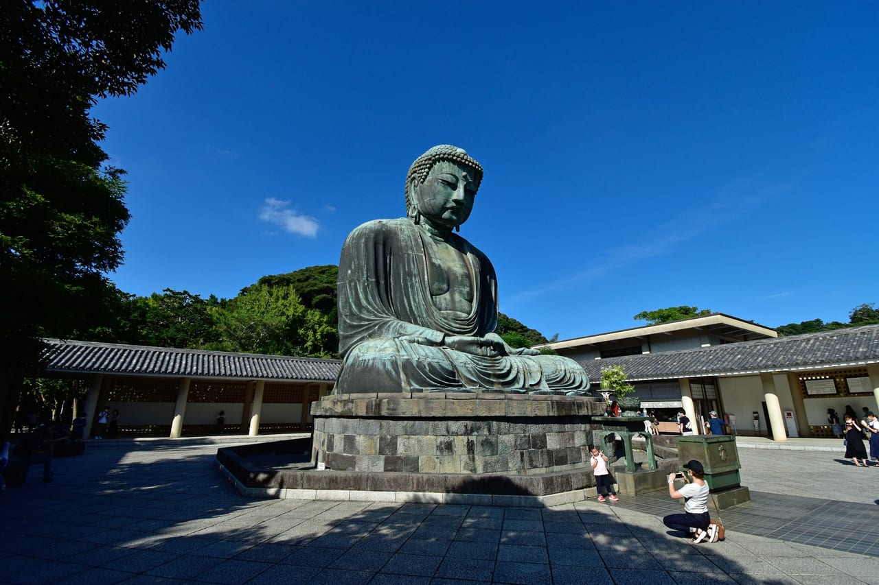 The Great Buddha of Kamakura, a giant weathered-bronze seated figure, sits outdoors against blue sky and spring cherry blossoms.