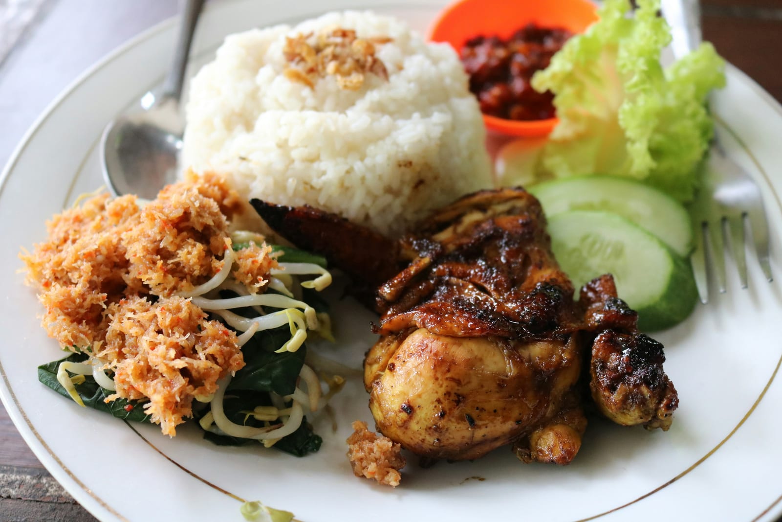 A plate of nasi campur with white rice, grilled chicken, vegetables, sambal, and shredded coconut.