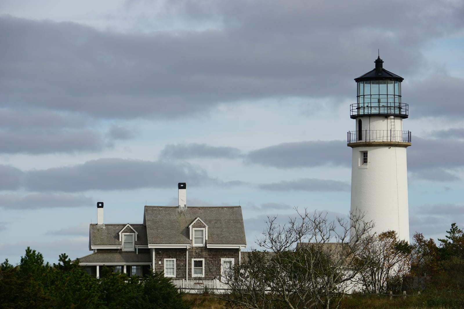 A shingled Cape Cod cottage beside a lighthouse under a moody sky