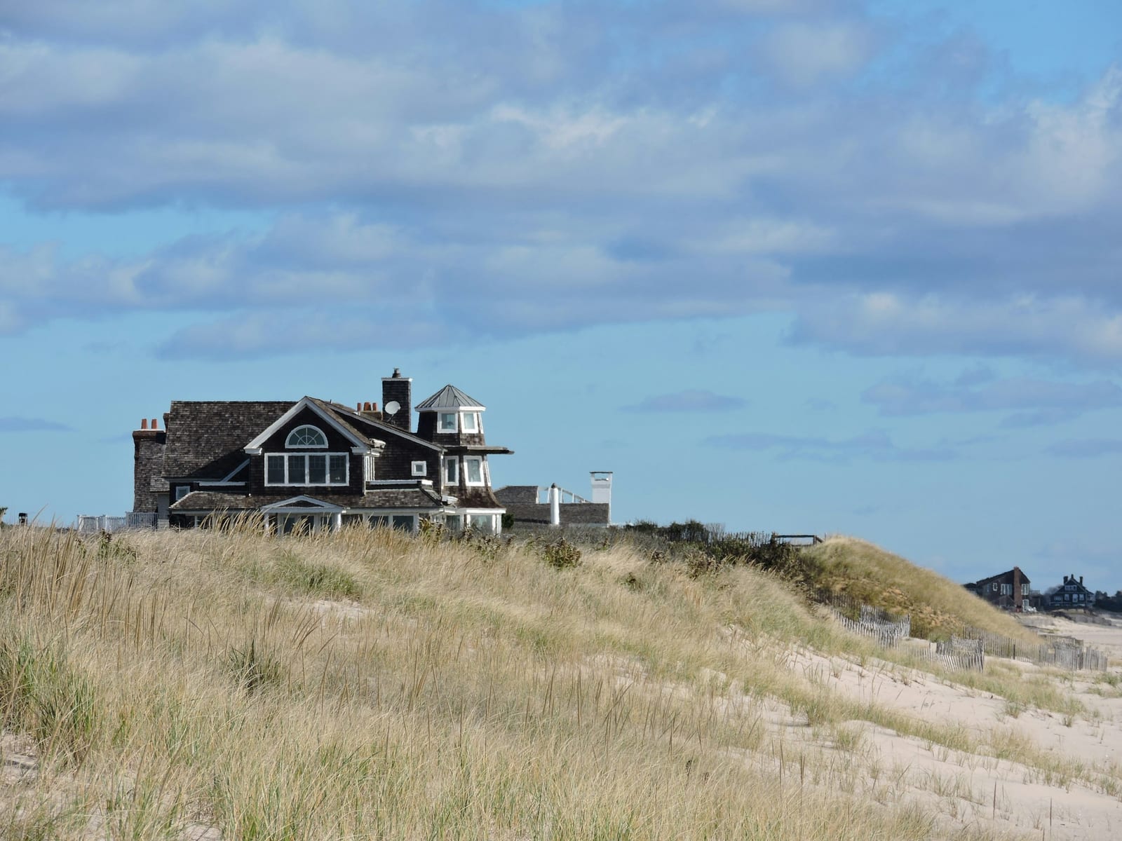 A shingled coastal house sits behind pale beach grass and dunes under a blue sky.