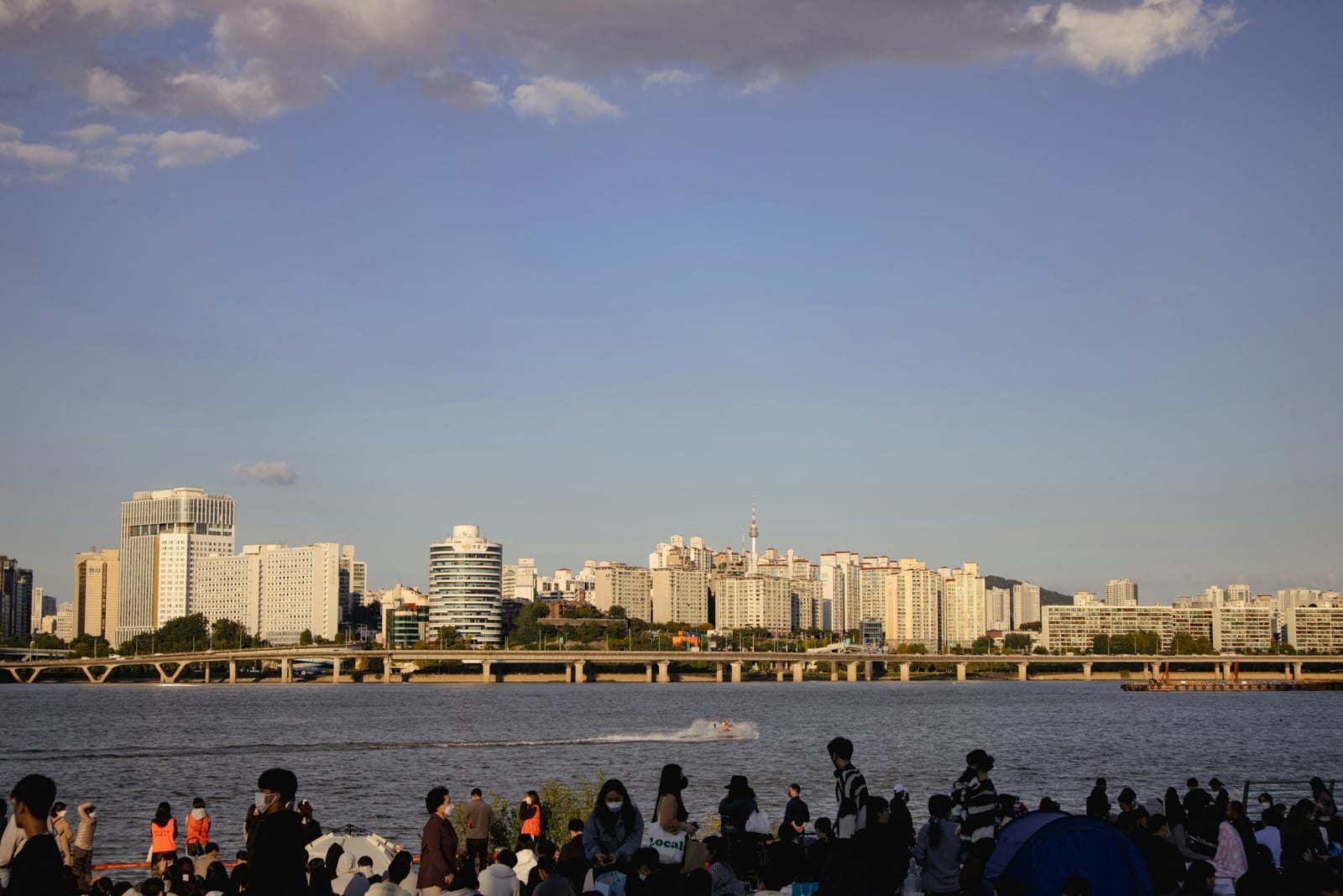 People gather along the Han River with Seoul’s skyline and a bridge beneath a clear evening sky.