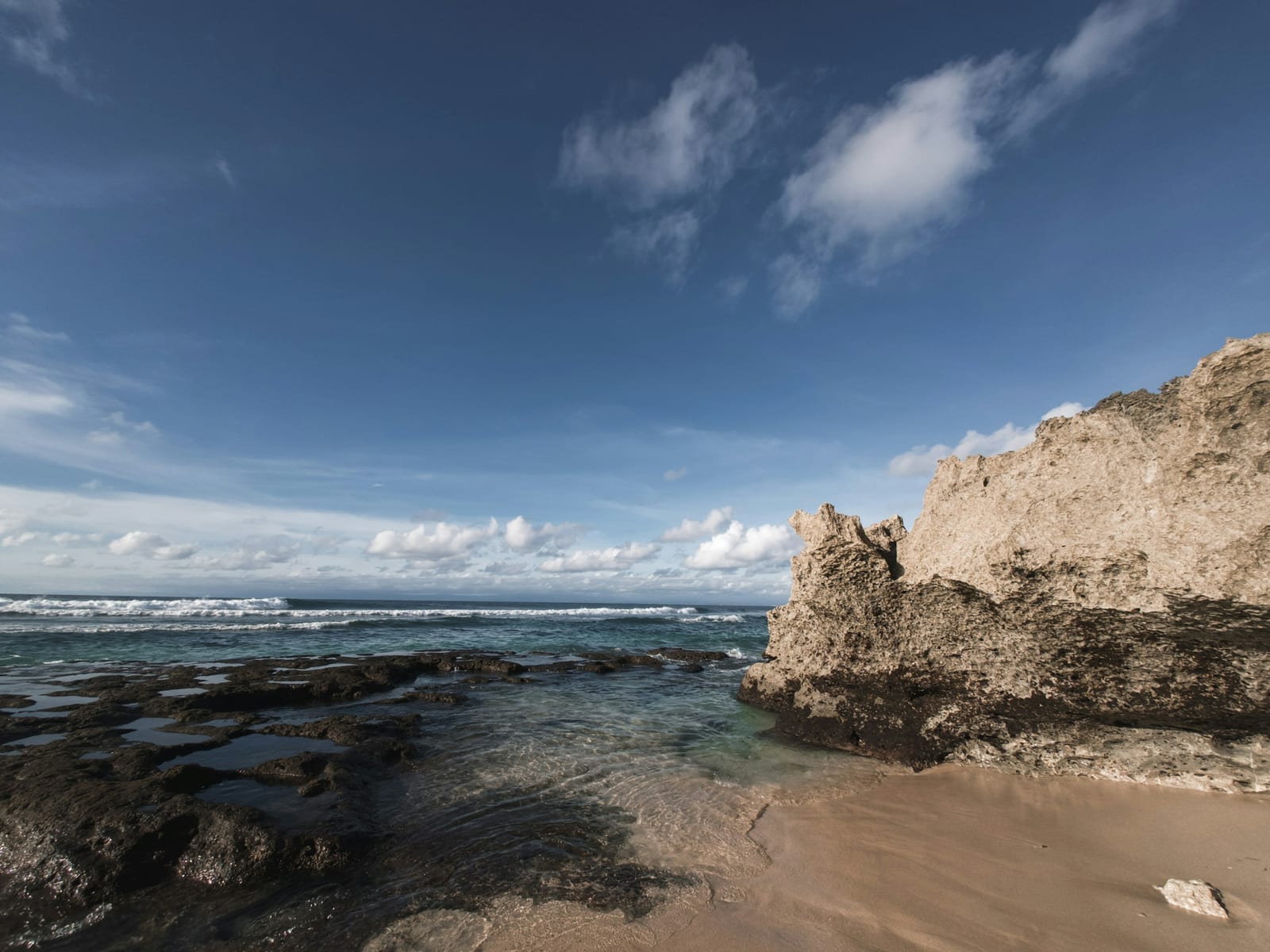 A sandy Uluwatu beach with rocky tide pools, surf, and a clear blue sky.