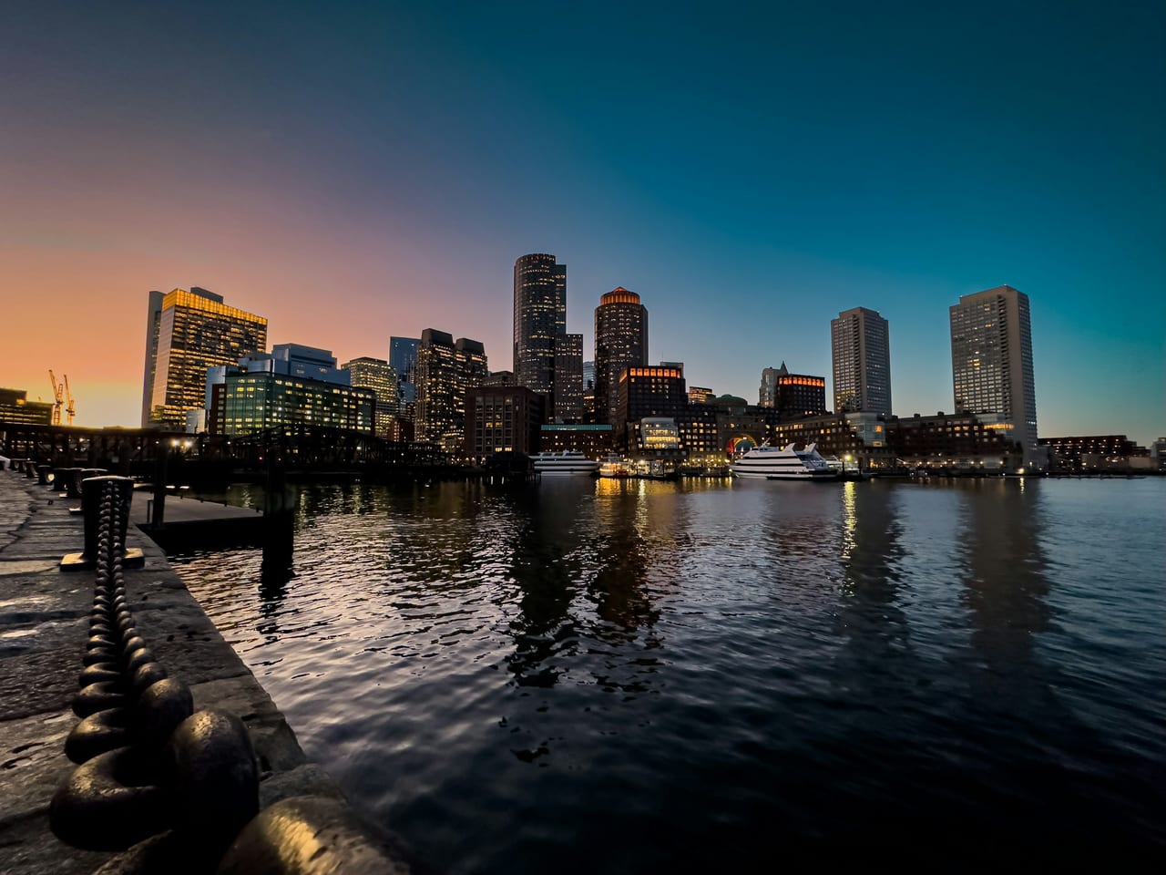 The Boston waterfront and Seaport skyline glow at twilight, reflected in the calm harbor water.