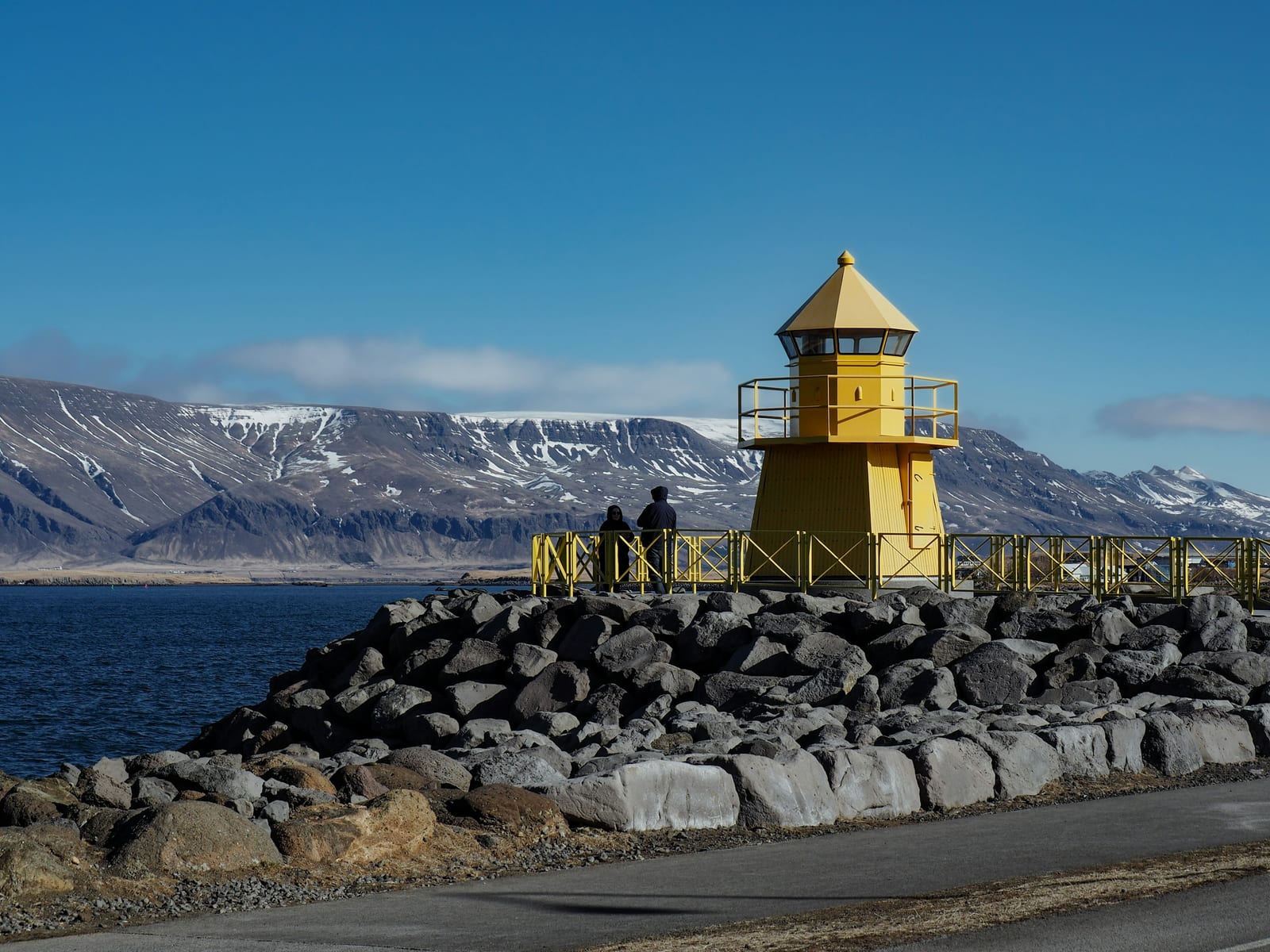 A yellow lighthouse stands on a rocky waterfront beside a road, with snow-streaked mountains across the water.
