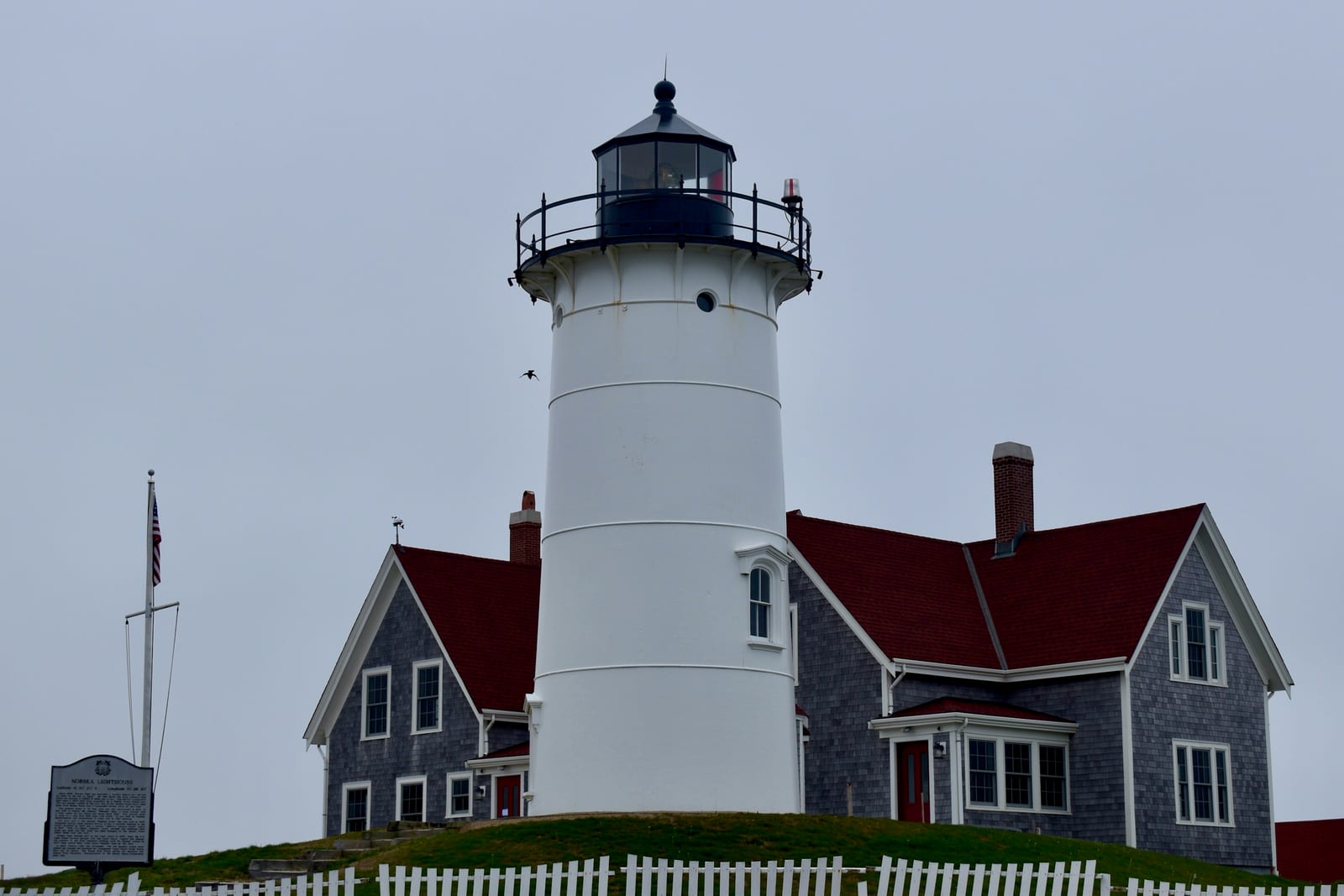 Nobska Light lighthouse with red-roofed keeper's house behind a white picket fence under an overcast sky in Falmouth.
