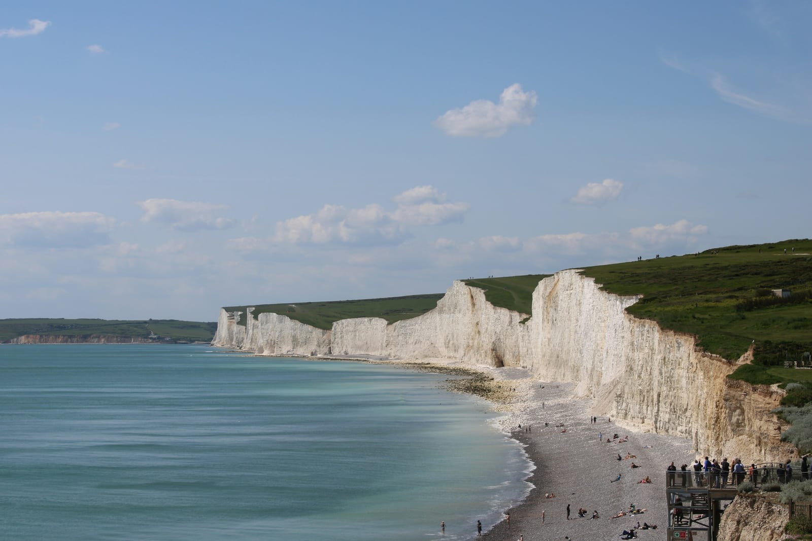 White coastal cliffs curve along a beach beside calm blue sea under a partly cloudy sky.