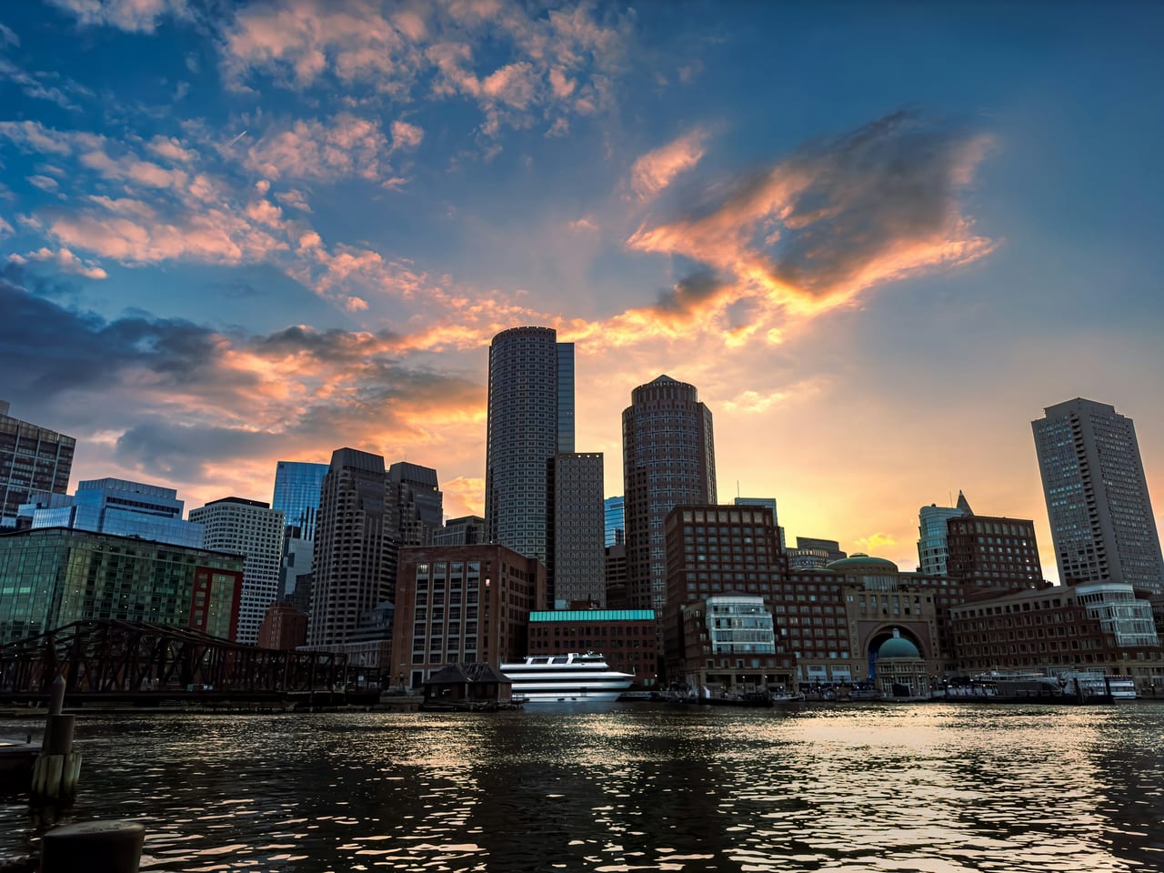 The Boston skyline reflects in the calm waters of the harbor at sunset, seen from the Seaport.