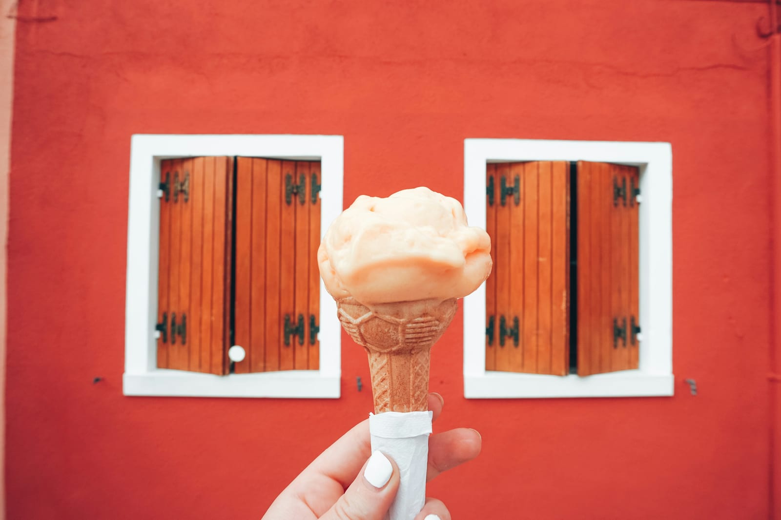 A hand holding a gelato cone in front of a bright red Roman building with wooden shutters.