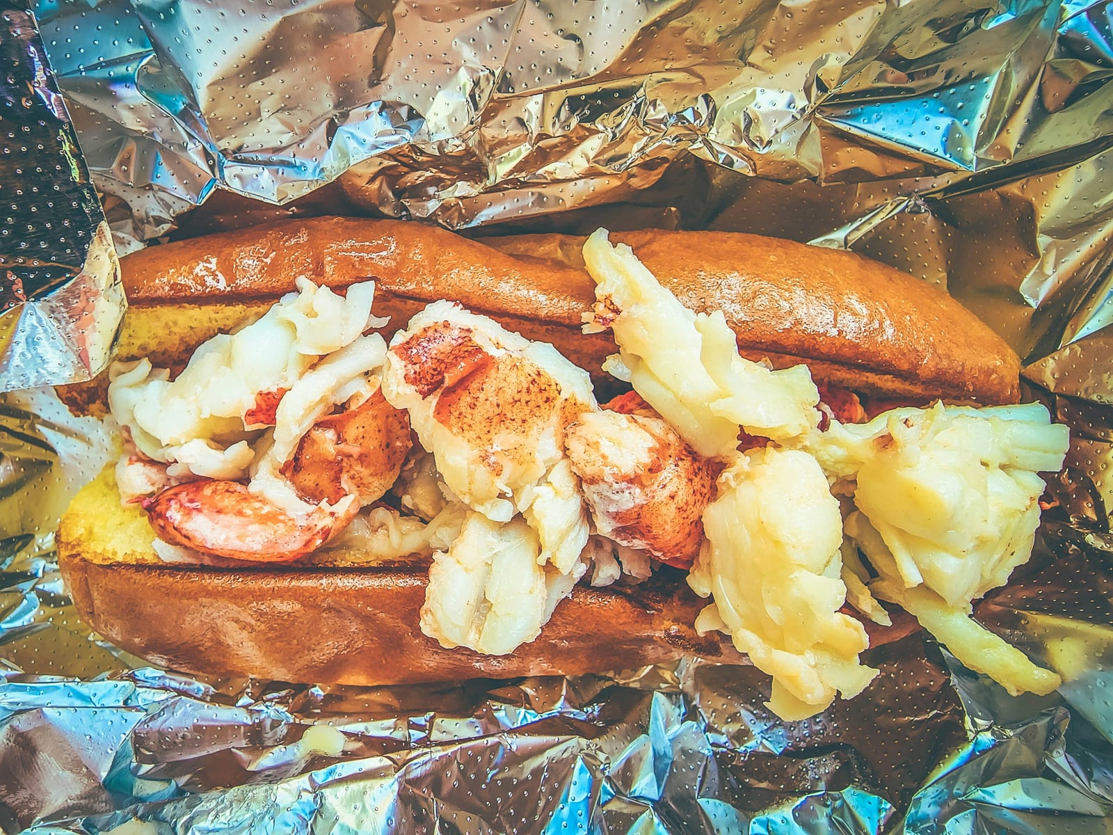 An overhead view of a lobster roll in a foil-lined tray, chunks of lobster meat piled into a split-top bun.