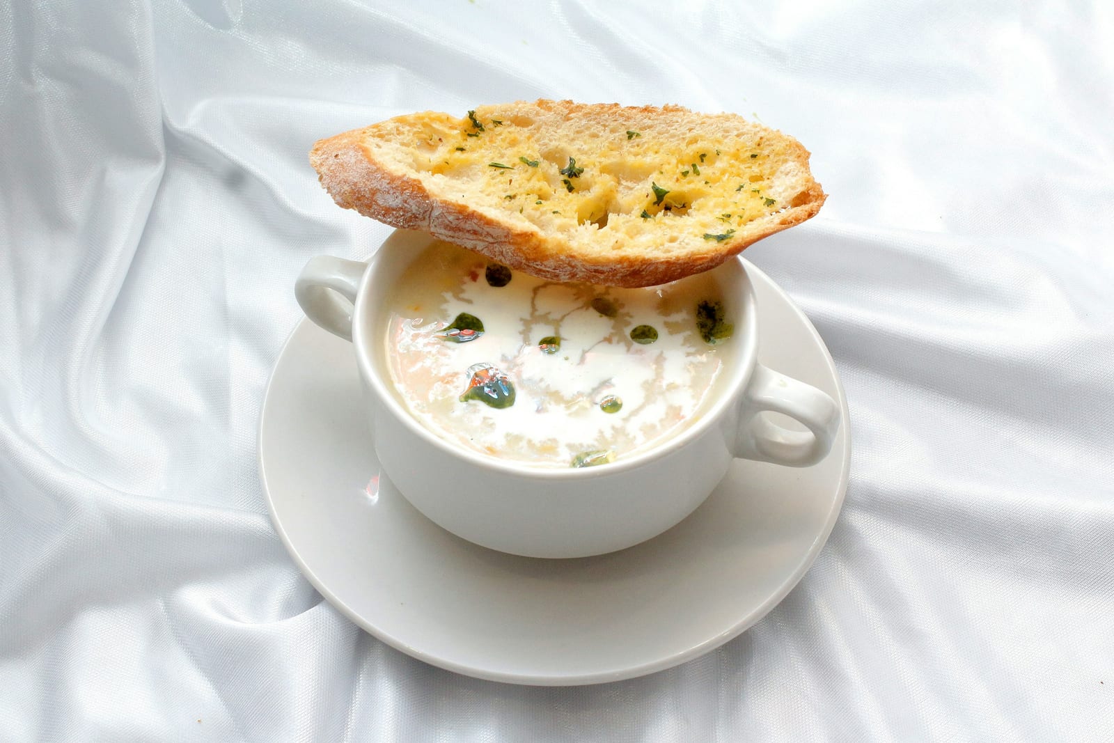 A white mug of creamy New England clam chowder topped with a garlic-bread crisp on a matching saucer.