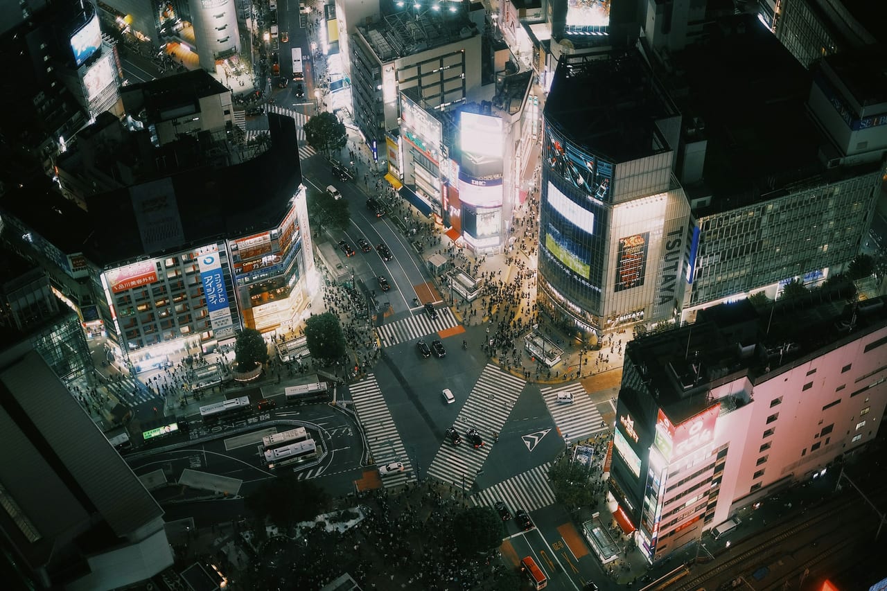 An aerial view of the Shibuya scramble crossing, with crowds streaming across its multi-directional crosswalks at dusk.