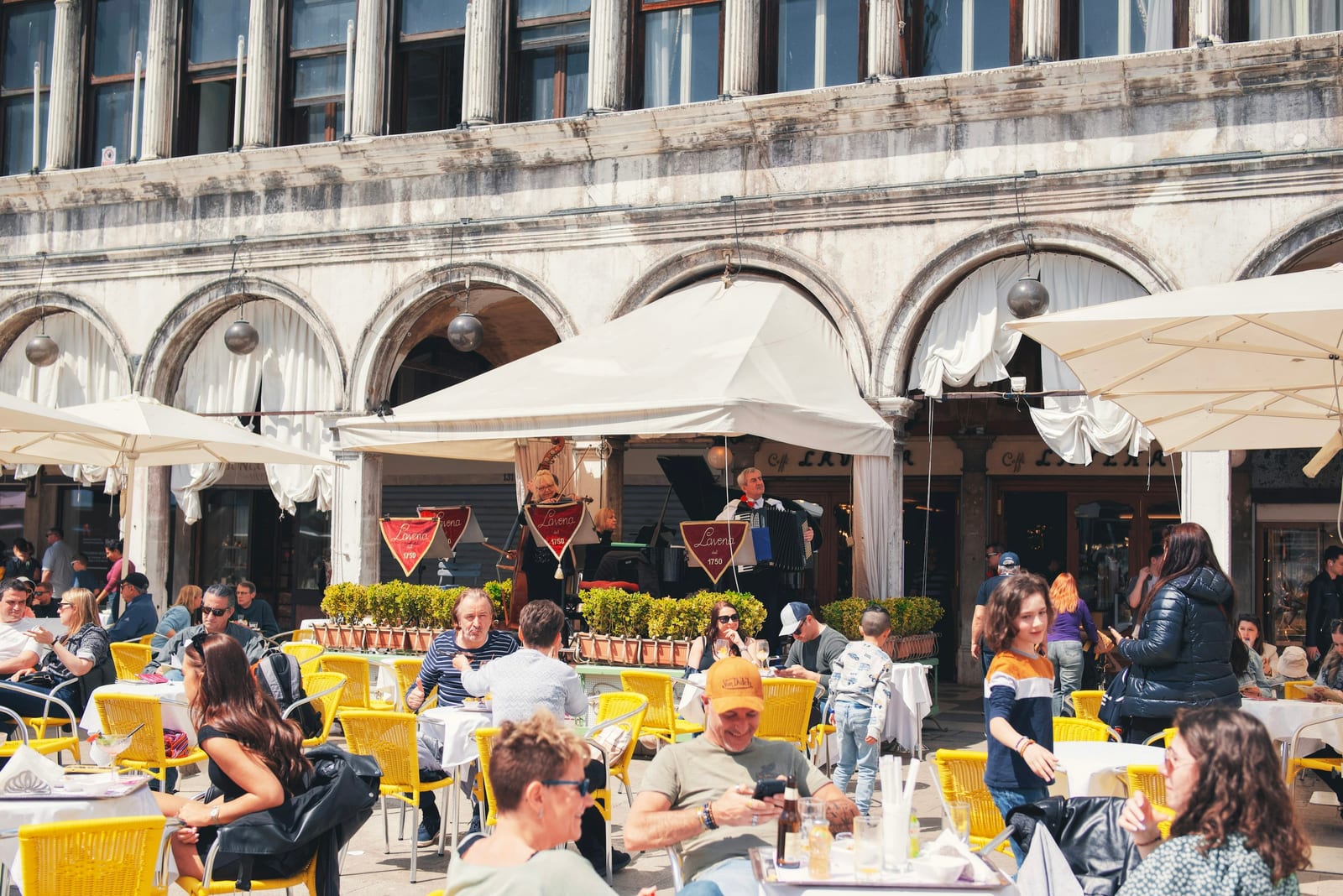 Tourists packed onto a busy Italian square at outdoor restaurant tables in front of a historic building.
