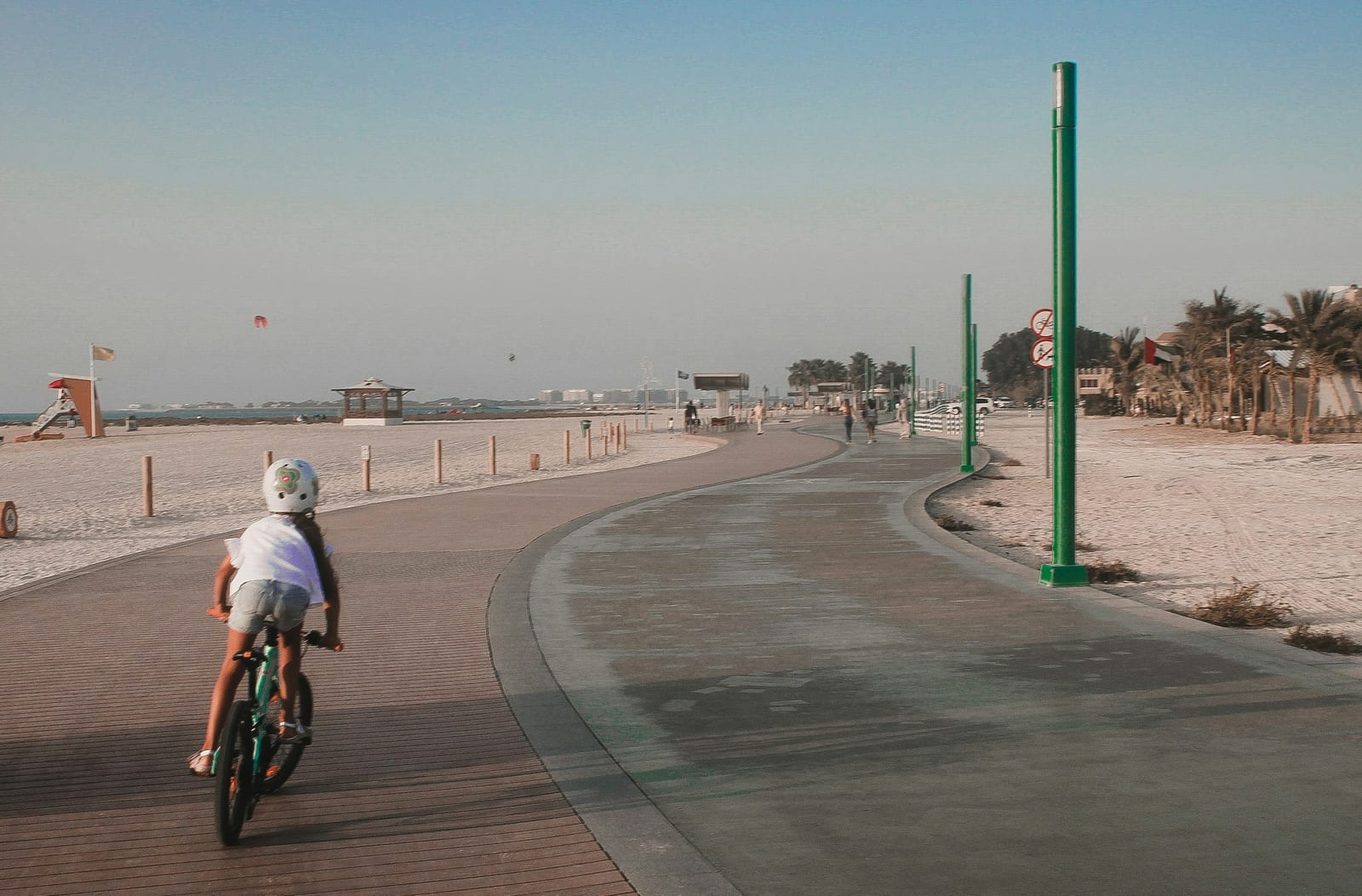 A child rides a bicycle along a paved path beside a sandy beach.