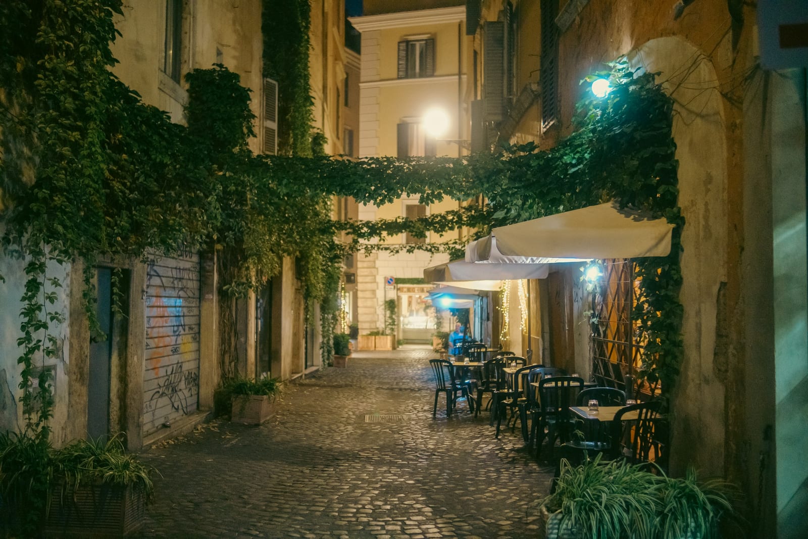 A cobblestone Trastevere lane with cafe tables set out at night.