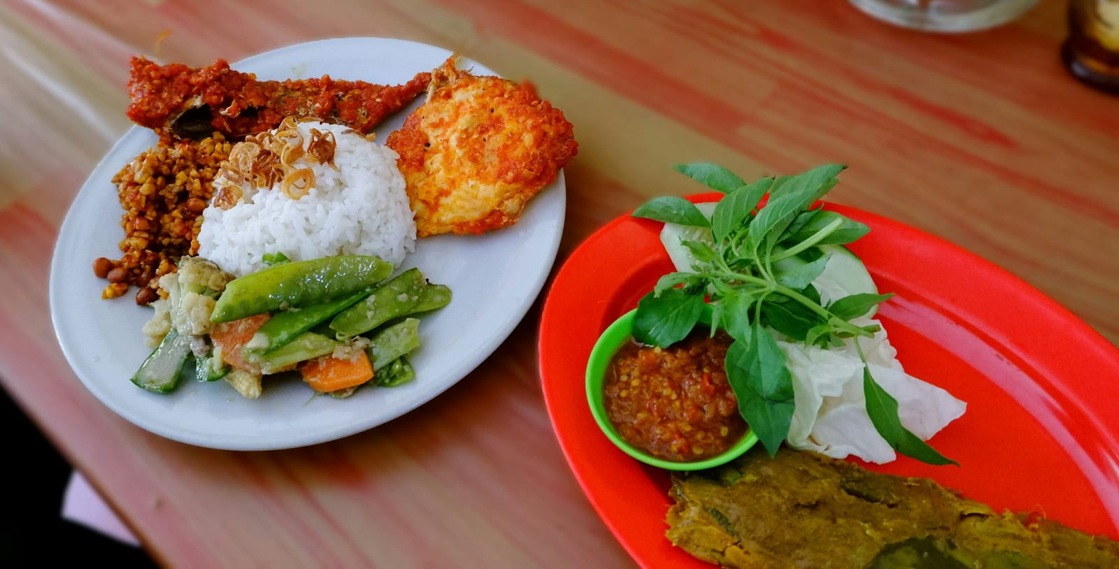 A colorful spread of rice, vegetables, fried snacks, and sambal served on plates and a red tray.