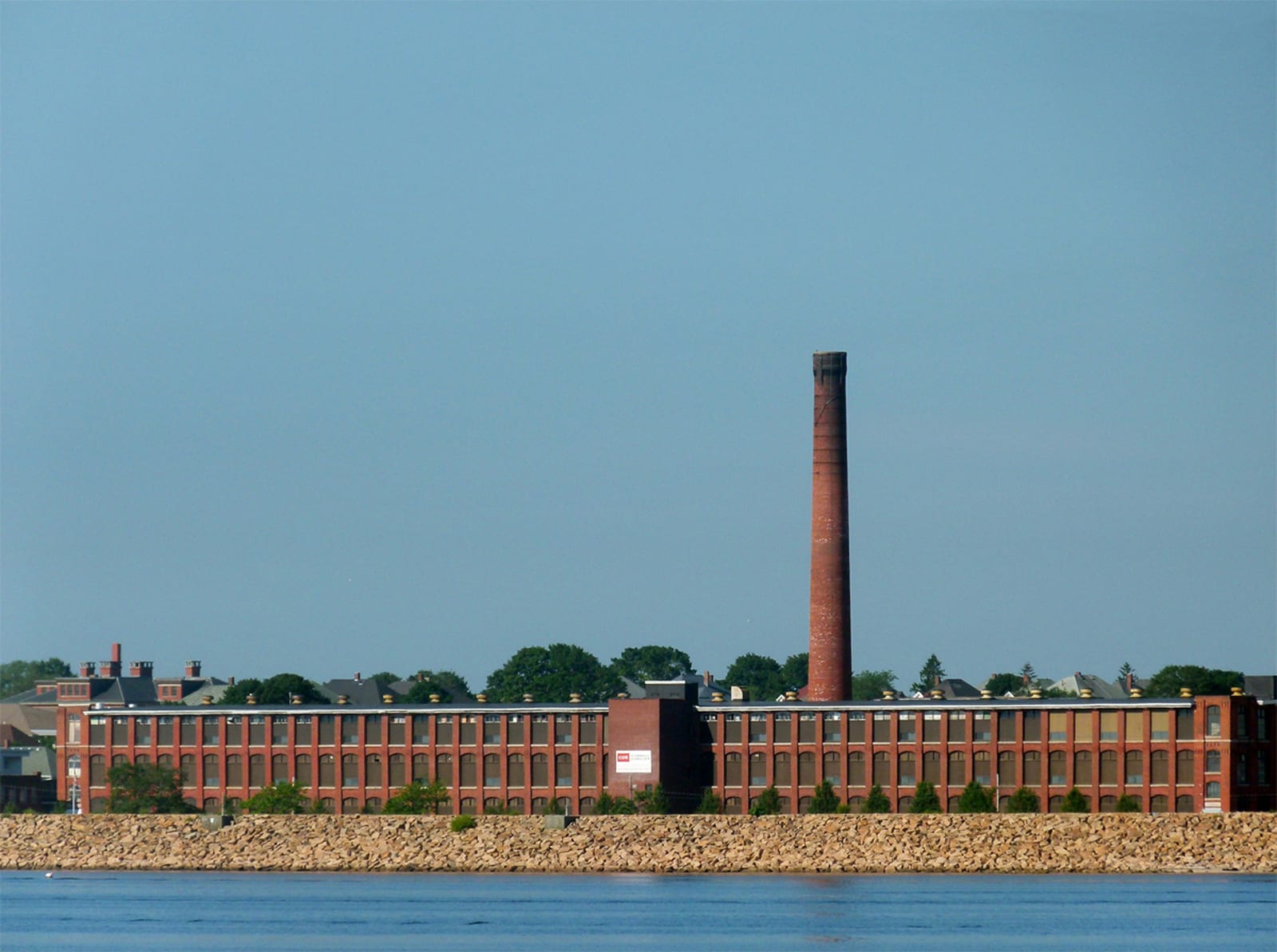 A long brick mill building with a tall smokestack sits along the waterfront under a clear blue sky.