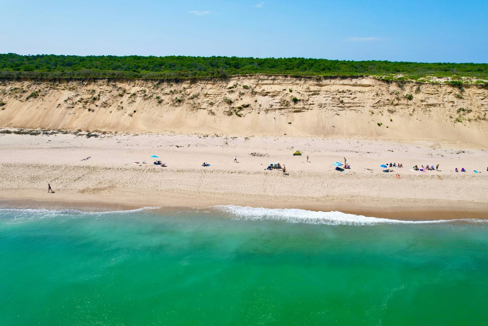 Aerial view of a sandy dune bluff meeting bright turquoise ocean water with a few beachgoers on the shore.