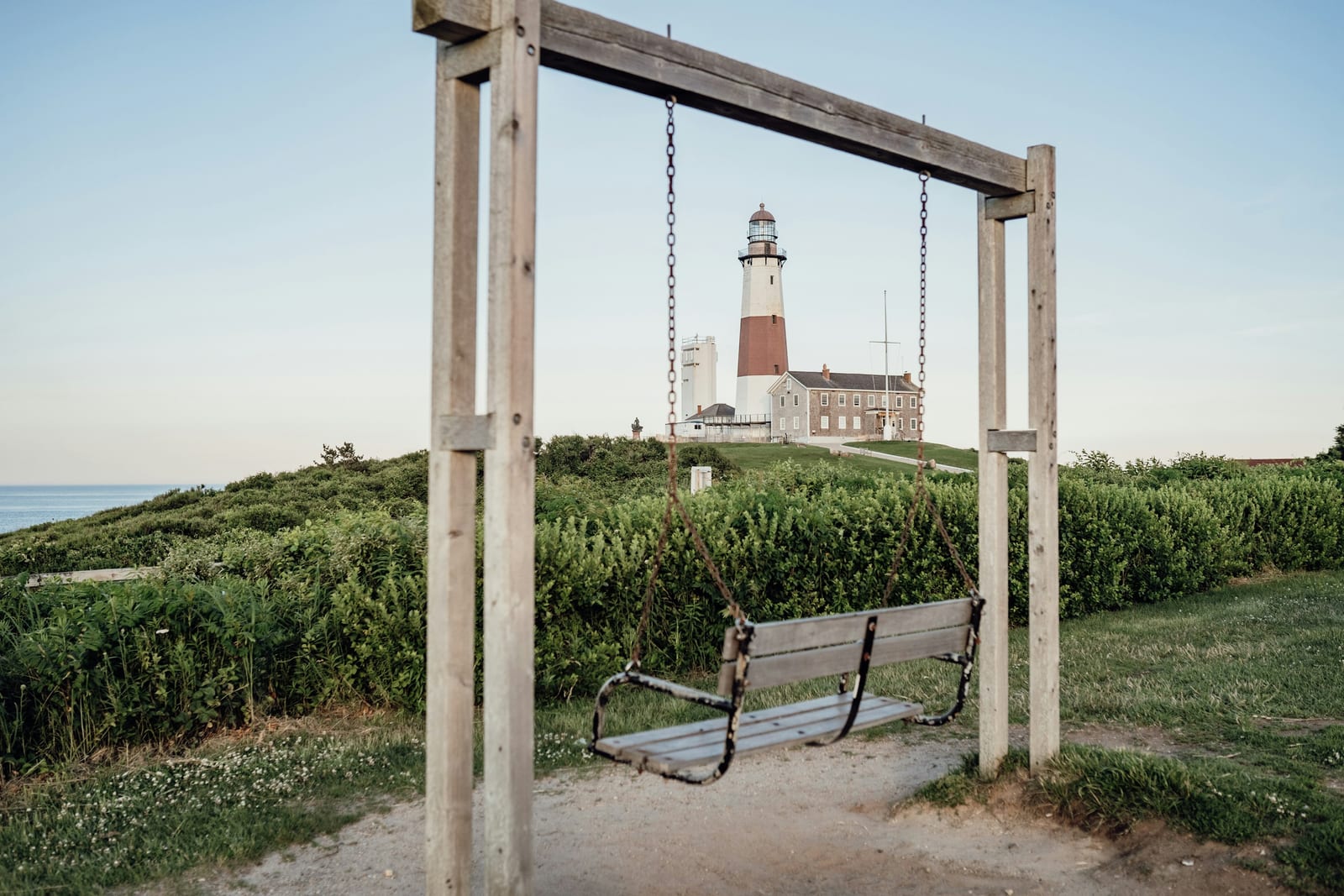 A lighthouse stands behind a wooden swing on a grassy coastal path.