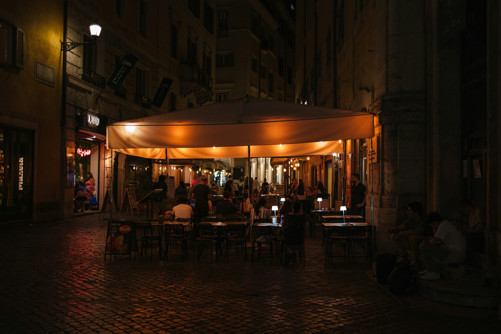 Diners under a lit canopy at an outdoor restaurant on a Roman cobblestone street at night.