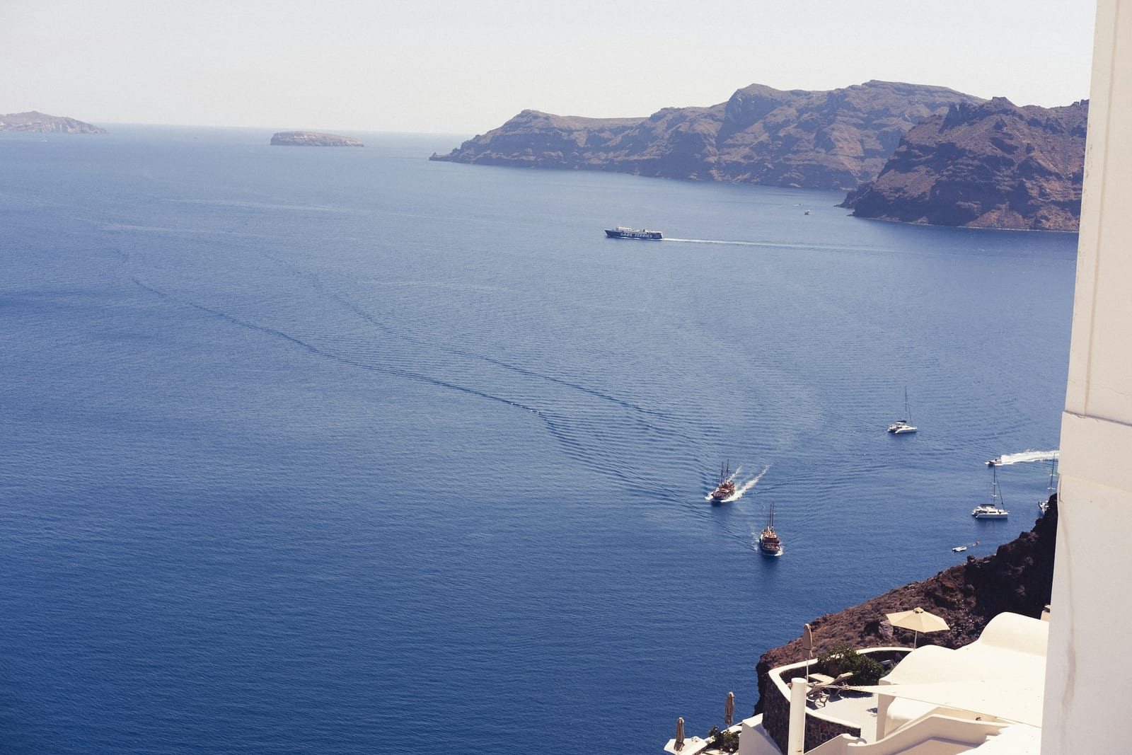 Several small boats cross deep blue water below a rocky island coastline and white seaside buildings.