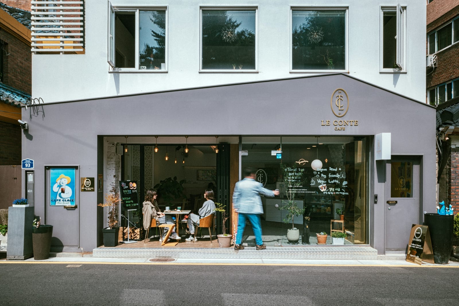 A bright Seoul cafe storefront opens onto the street, with customers seated inside and drinks displayed near the counter.