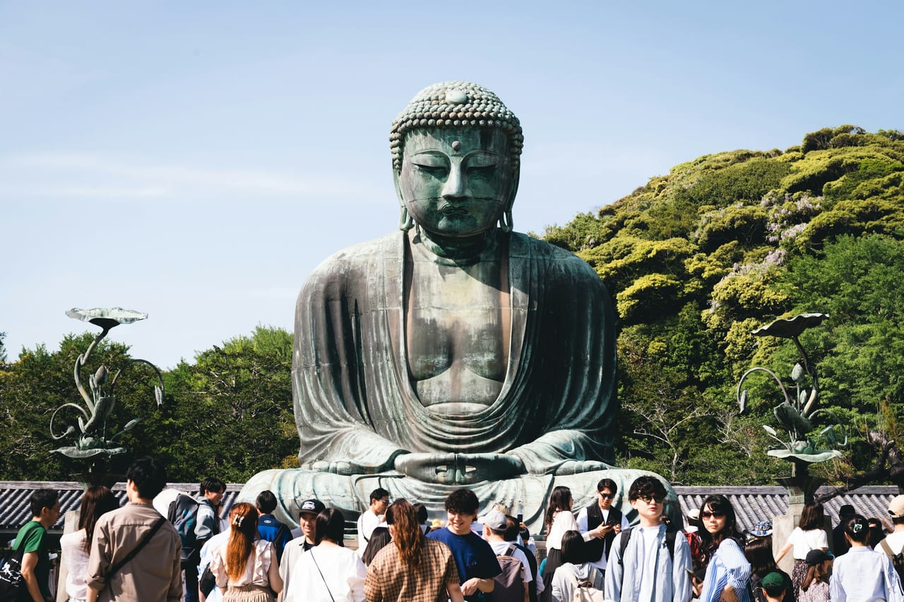 The Great Buddha of Kamakura, a giant weathered-bronze seated figure, sits outdoors against blue sky and spring cherry blossoms.