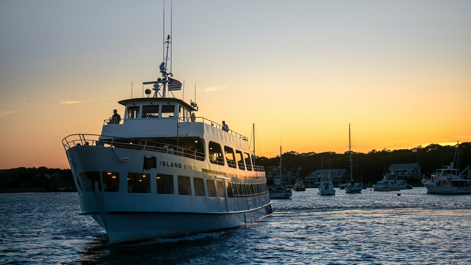 A passenger ferry moves into the harbor at sunset with boats and shoreline homes behind it.