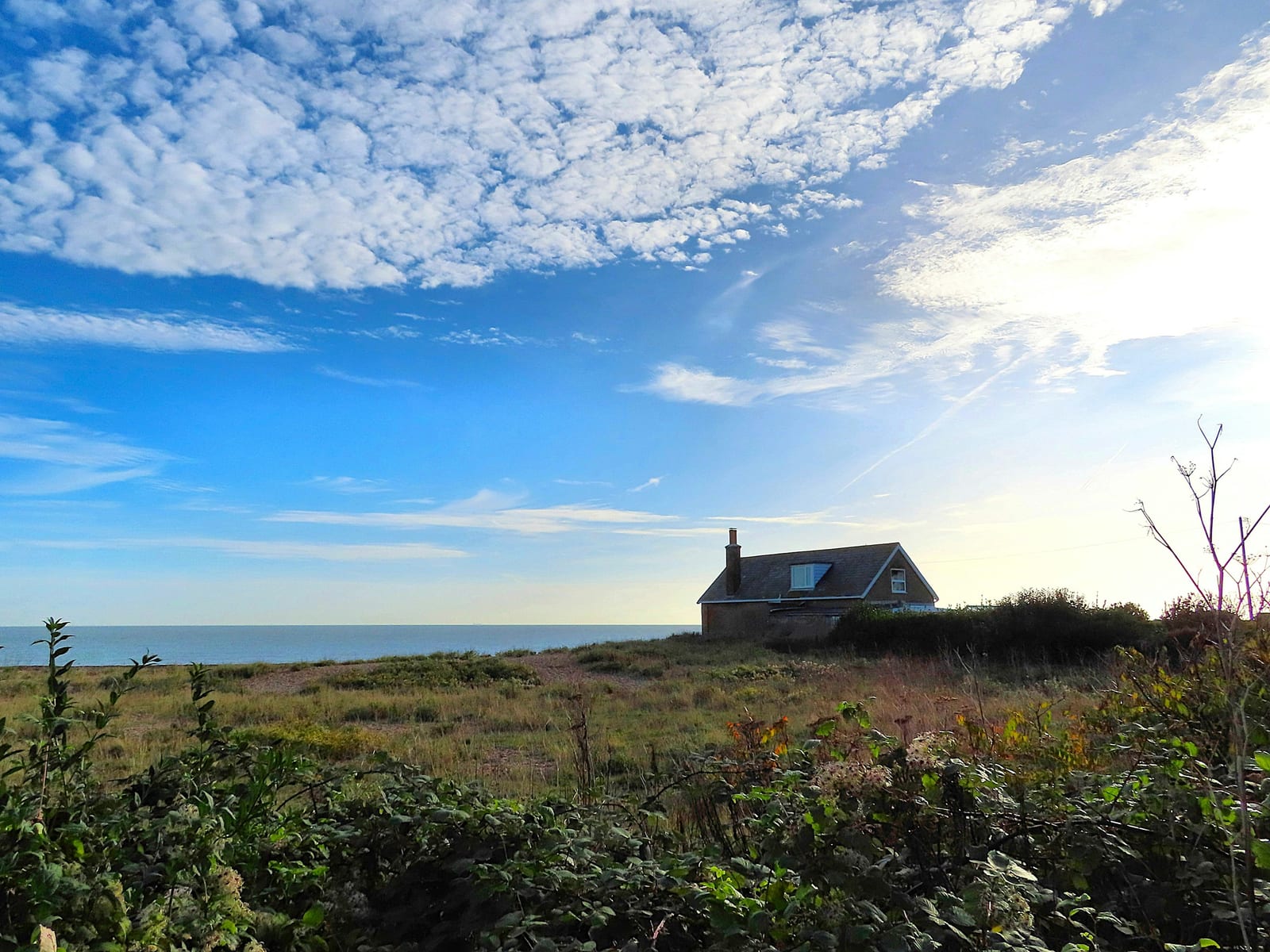 A small cottage overlooks the ocean from a grassy bluff dotted with autumn shrubs under a sunny sky.