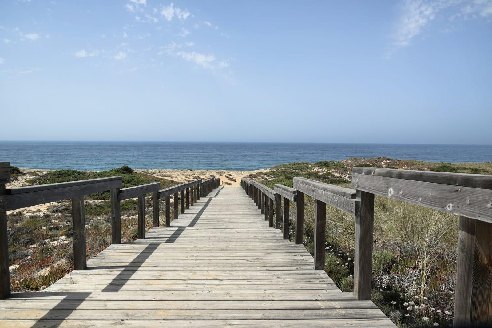 A wooden boardwalk crosses beach dunes toward the ocean under a bright blue sky.