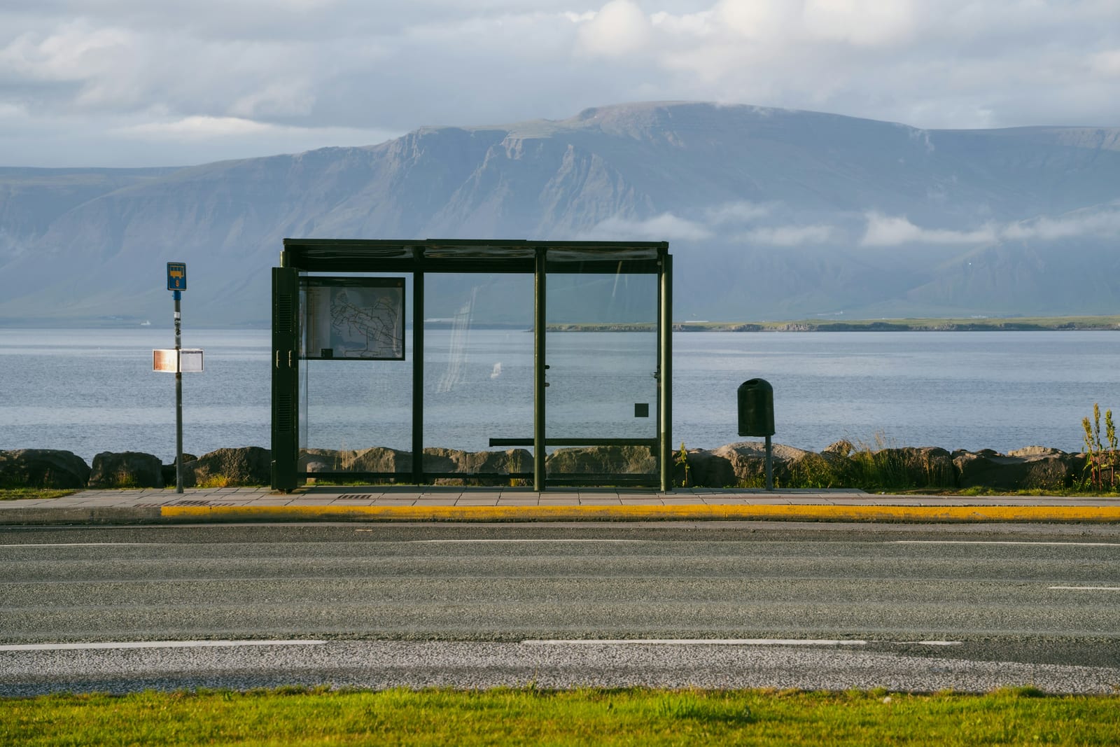 An empty bus shelter stands beside a road with a lake and broad Icelandic mountains behind it.