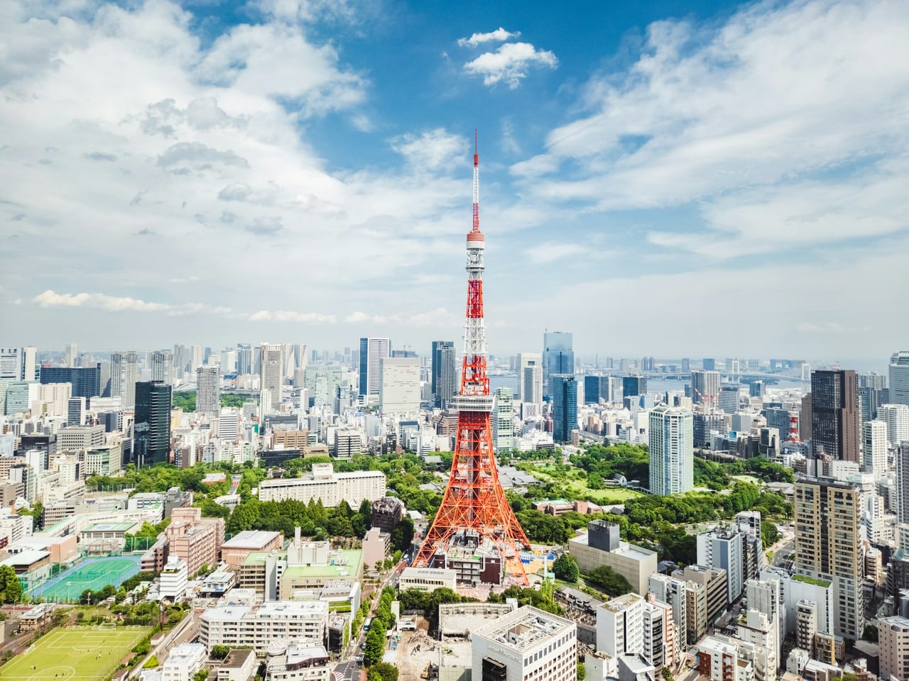The red-and-white Tokyo Tower stands above a sprawling daytime skyline under a clear blue sky.
