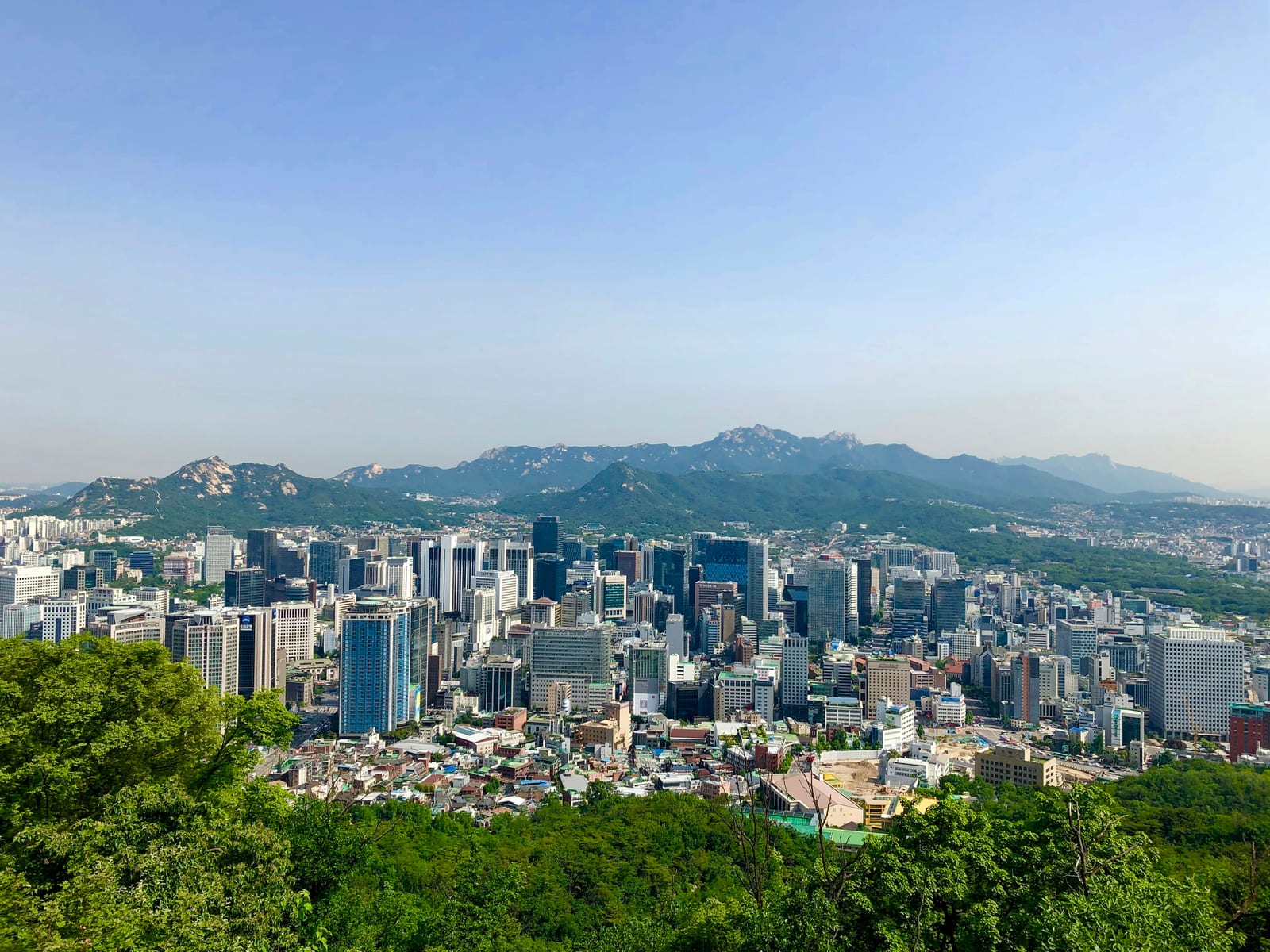A bright view of Seoul's skyline with a wooded hill in the foreground and mountains behind.