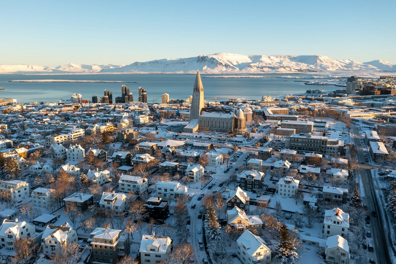 A bright aerial view of snow-covered Reykjavik with Hallgrimskirkja, the harbor, and mountains beyond.