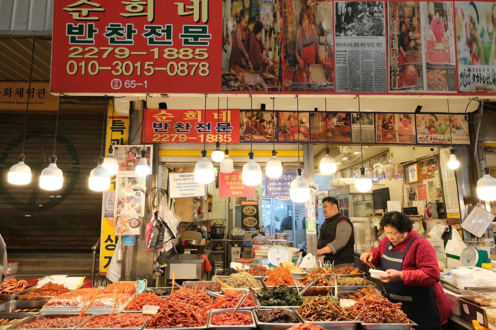 A brightly lit Korean market stall displays trays of prepared seafood beneath Korean signs, with vendors working behind the counter.