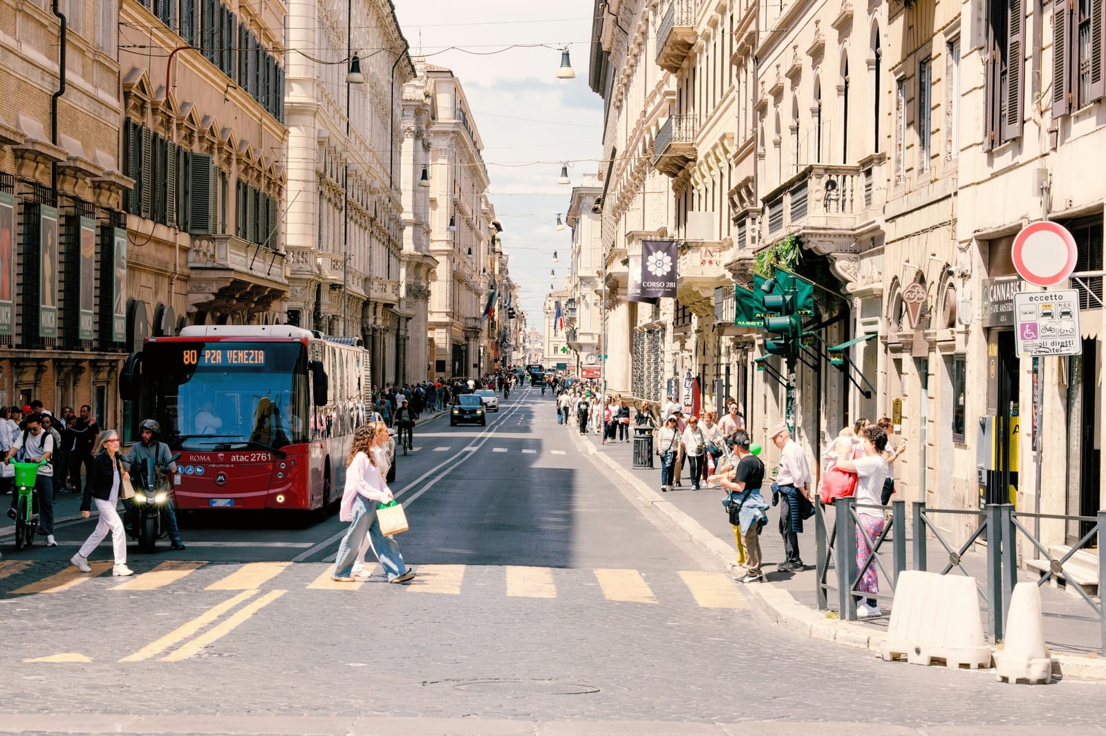A red ATAC city bus on Via del Corso in central Rome as people cross the street on a sunny day.