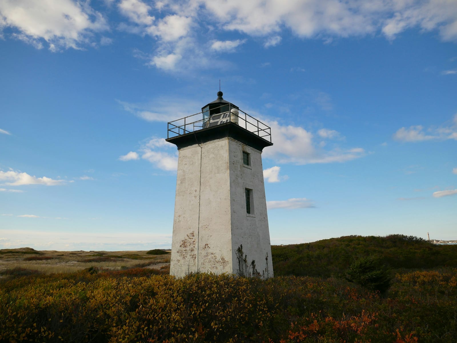 A white lighthouse tower stands among reddish-brown autumn shrubs under a clear blue sky with scattered clouds.