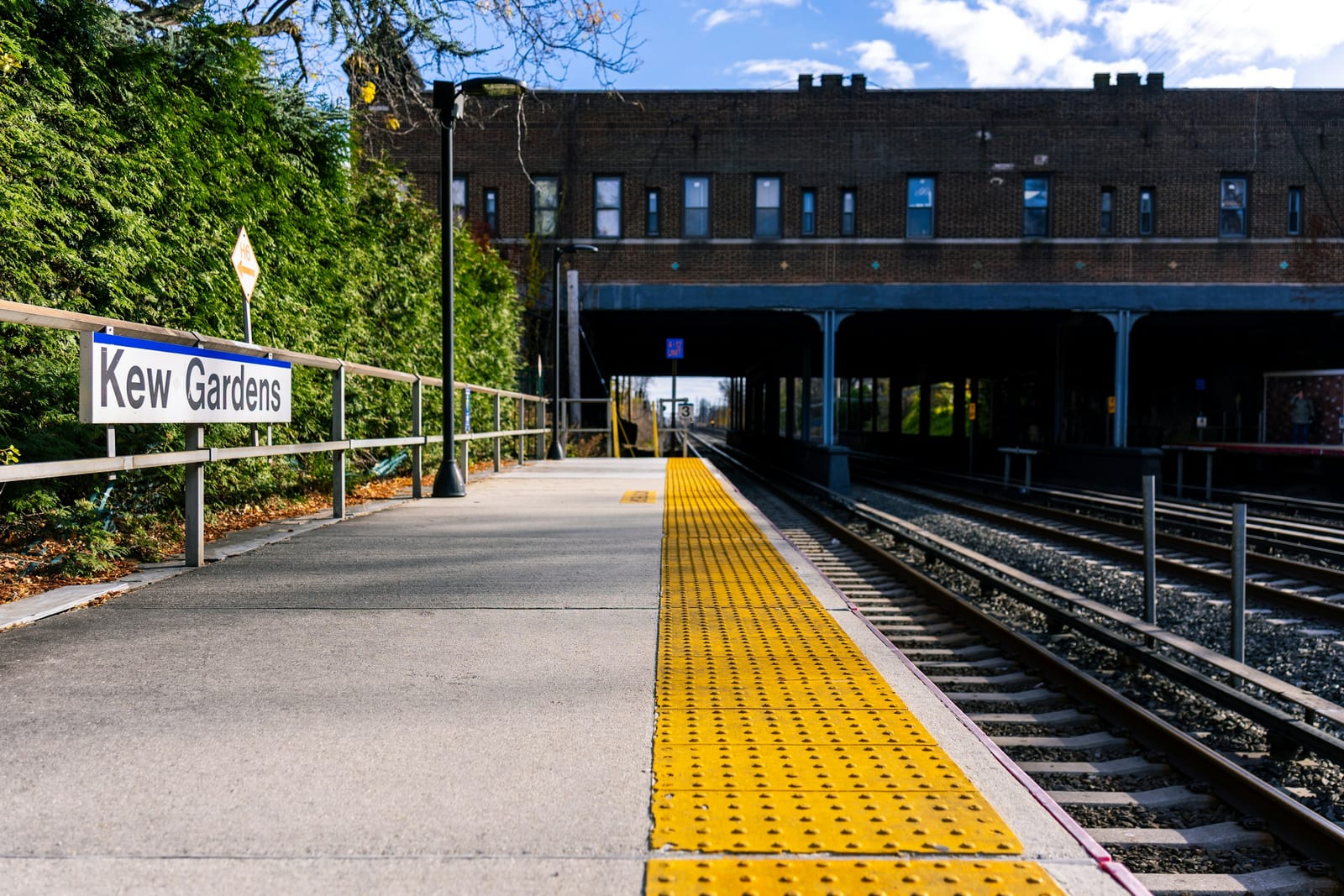A sunny Kew Gardens train station platform with yellow tactile paving, tracks, and a station building.