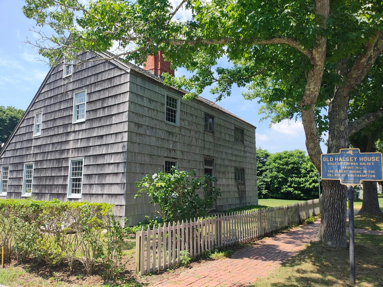 A historic shingled house beside a tree-lined path and picket fence.