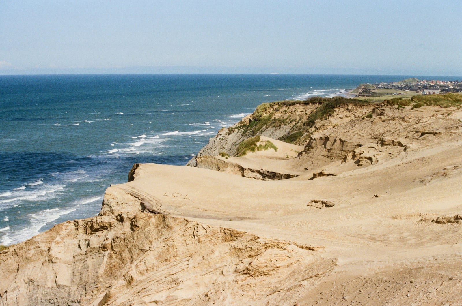 Sandy cliffs overlook a blue ocean with distant buildings.