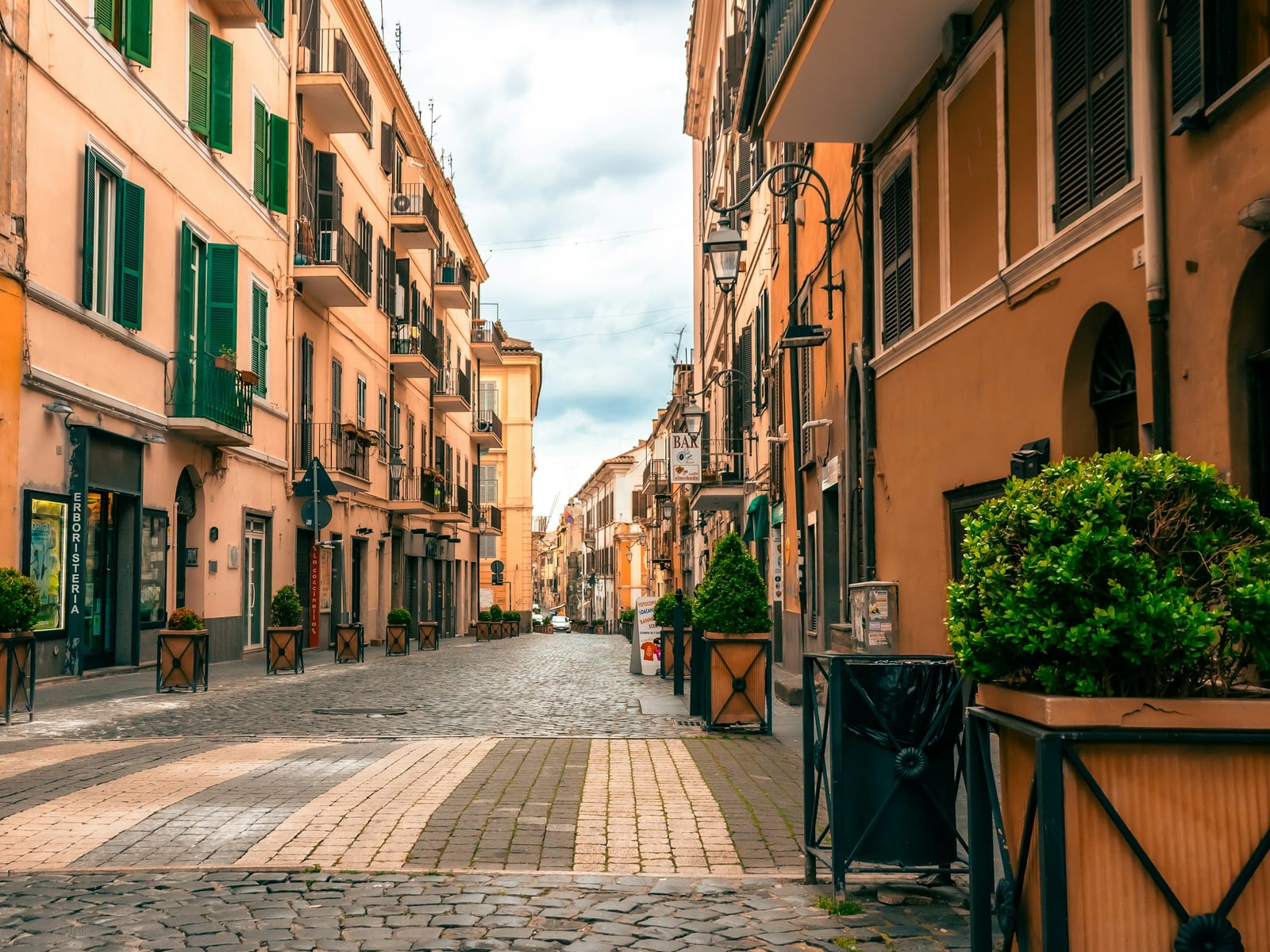 A quiet residential lane in Rome with ochre buildings, green shutters, potted plants, and cobblestones.