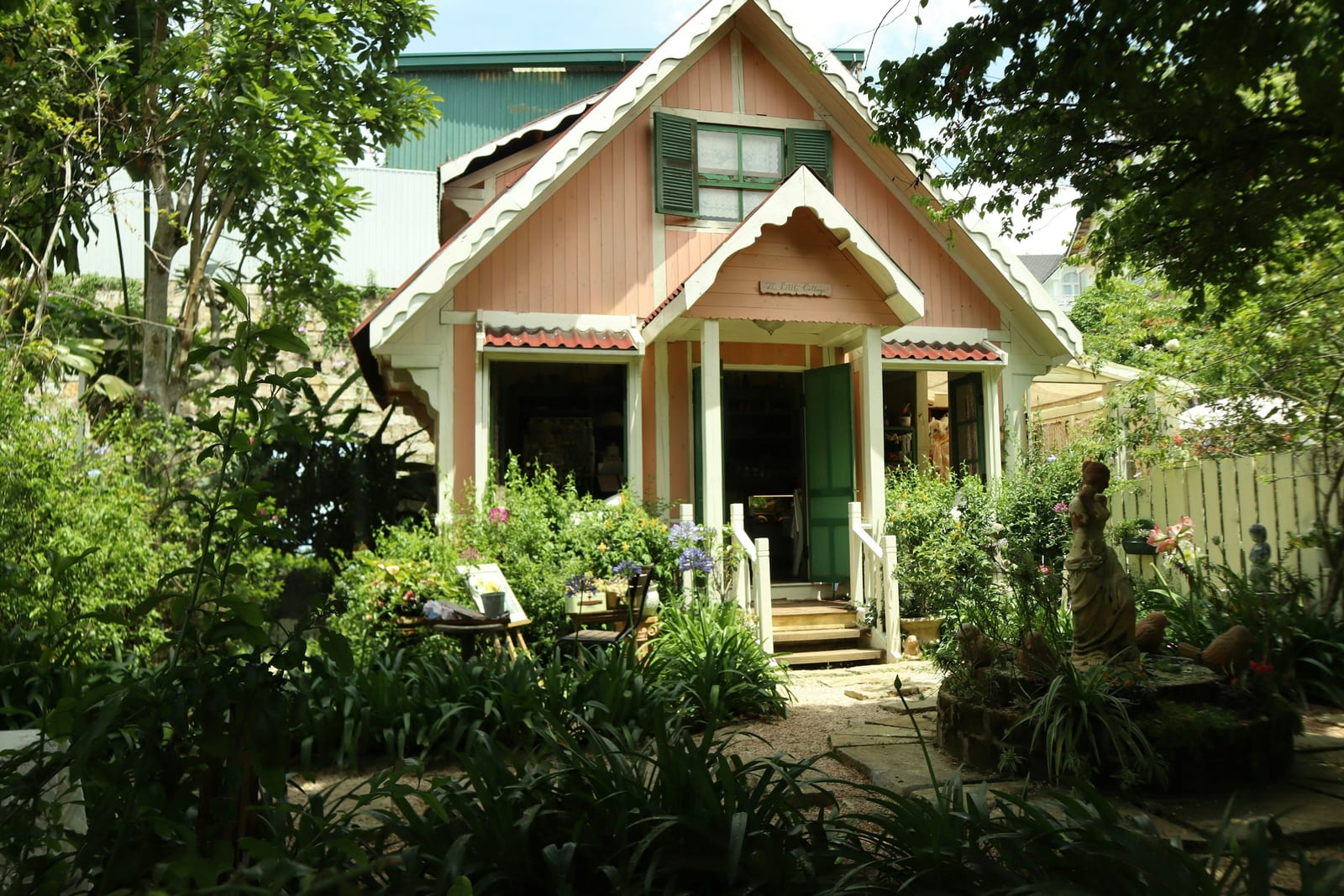 A small, light-colored guesthouse with a peaked porch stands among leafy garden plants.