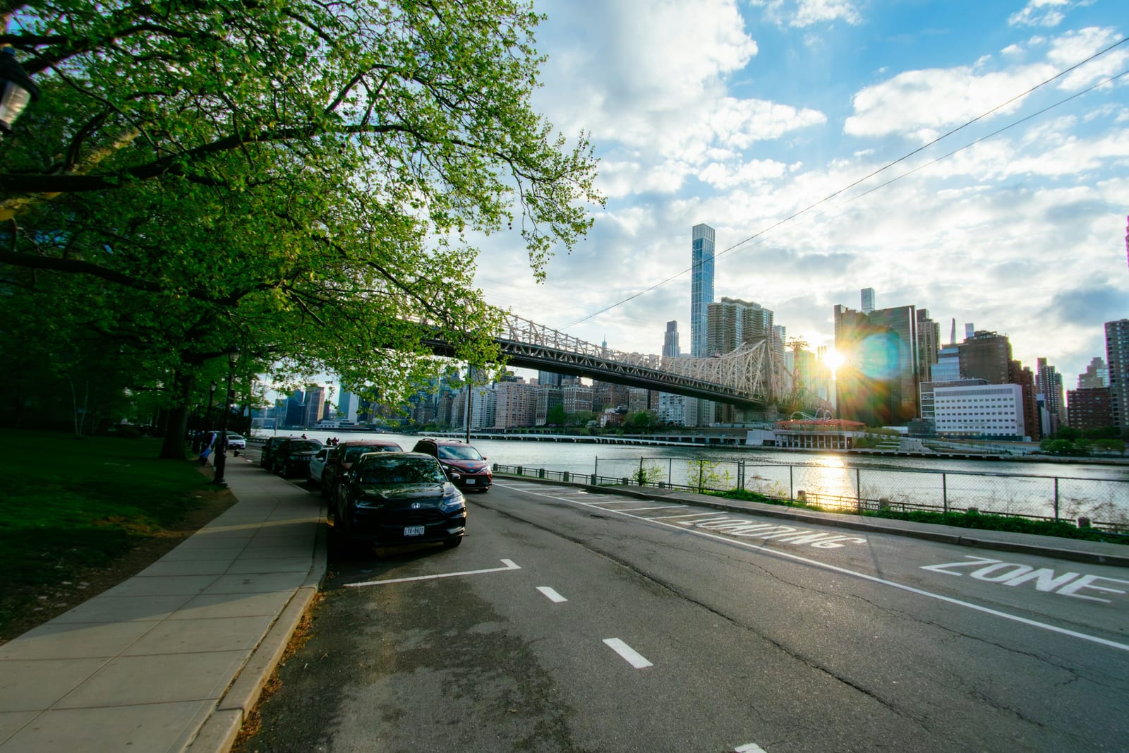 A sunlit city road runs beside a river with a bridge and skyline in the background.